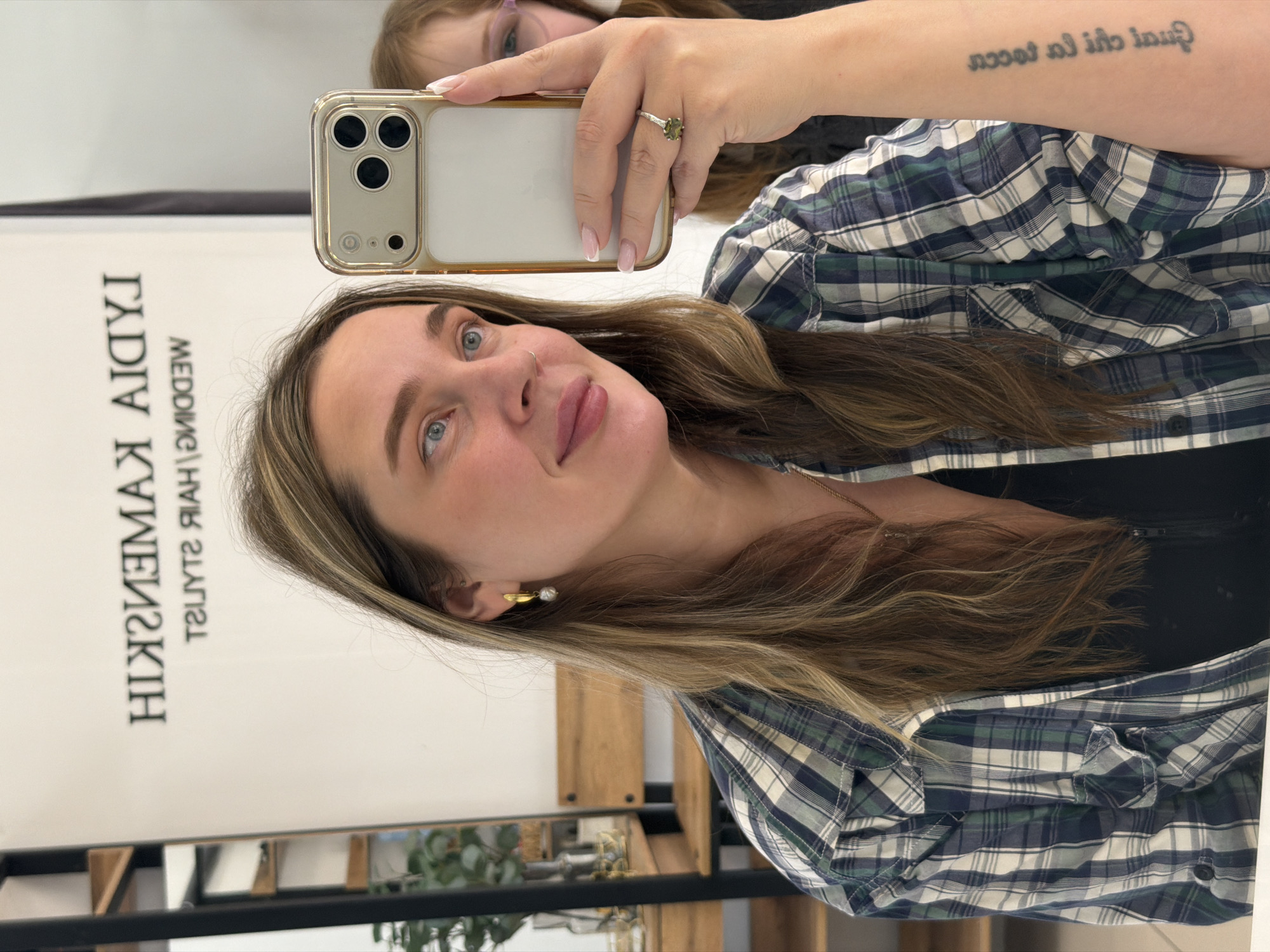 A woman is depicted taking a mirror selfie in an indoor setting, likely a salon or beauty studio. She holds a light-colored iPhone, equipped with a multi-lens camera system and housed in a clear or light-gold case, up towards the mirror to capture her reflection. Her right hand, adorned with a ring featuring a green stone and manicured with a French tip style, is visible holding the phone.
The woman has long, light brown hair with blonde highlights, blue eyes, and a light pink lip color. She wears a plaid button-up shirt in shades of blue, green, and white. A small nose piercing is visible, and she wears subtle gold-toned earrings. Her expression is a soft, upward gaze towards the camera, with a slight smile. On her right forearm, a tattoo is visible, reading "Qual la cio la tocc" (reversed).
Reflected in the upper left portion of the mirror, the partial face of a child wearing glasses can be seen. The background reflected in the mirror features a white wall with reversed black text, which appears to include "IADIAW" (part of "WEDDING" reversed), "KIMSHIH" or "HISHEMA" (possibly a name, reversed), and "TSUYTS RYAH" ("HAIR STYLIST" reversed). Below this text, a wooden shelving unit with small potted plants or decorative items is also reflected. The lighting is bright and even, consistent with an indoor daytime setting.