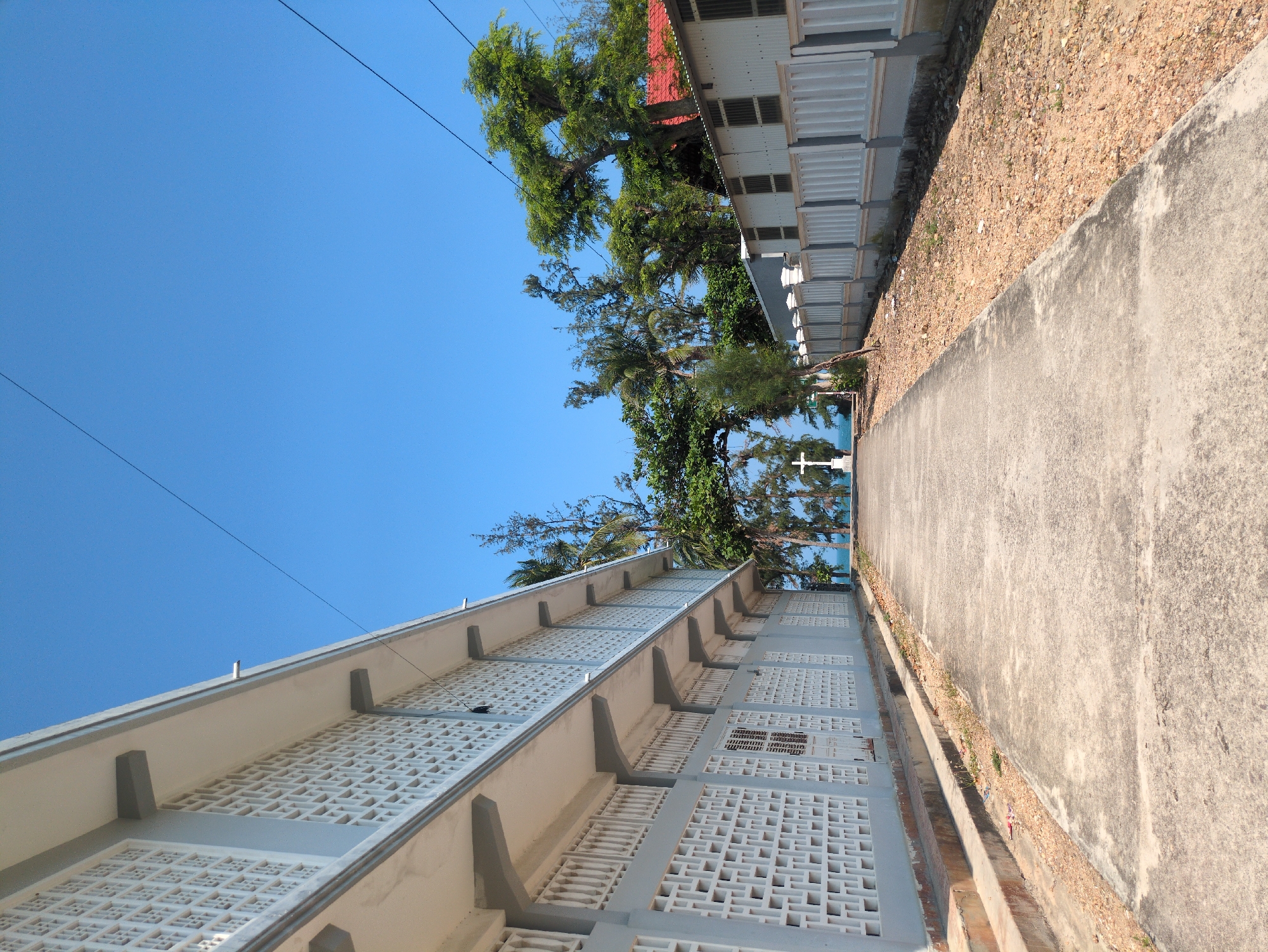 The image depicts a concrete path running between two buildings under a clear blue sky in South Quy Nhon Ward, Vietnam. The path angles towards the horizon, meeting the sea in the distance. On the left, a multi-story building with a repeating geometric pattern on the exterior wall is visible. On the right, a lower building with a red roof section can be seen among trees. Power lines stretch across the sky above. A small white cross is visible near the horizon.