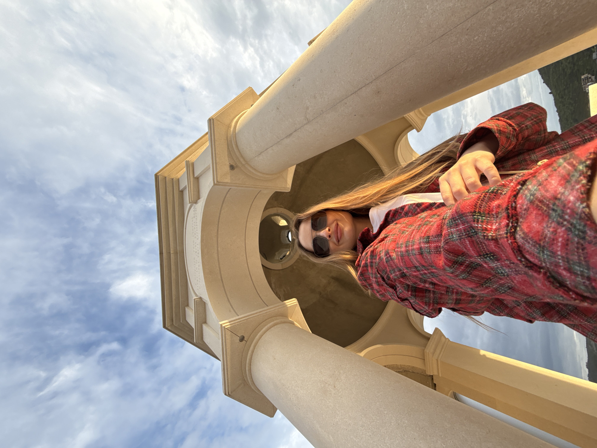A low-angle, tilted shot looking upward features a woman in sunglasses and a red plaid jacket standing under a stone structure with columns and a rounded top. The architecture is off-white or light beige. The sky is visible through openings in the structure and is partly cloudy. The location is Đuraševići, Montenegro.