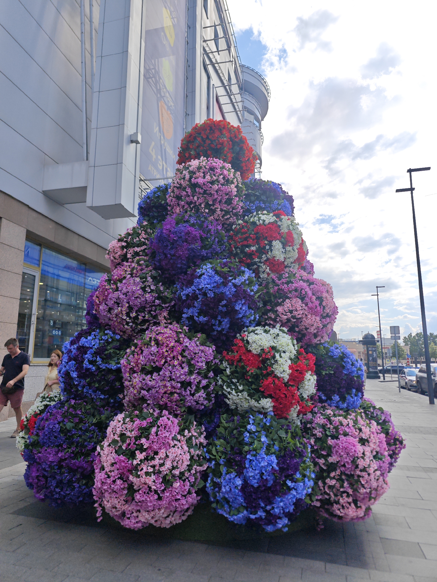 The image shows a large, cone-shaped floral arrangement displayed outdoors on a city sidewalk in front of a modern building. The floral arrangement is densely packed with small, colorful flowers in shades of pink, purple, blue, red, and white. The building behind the arrangement appears to be a commercial establishment with large windows and signage. Two individuals are visible on the sidewalk near the building: a man in a dark polo shirt and shorts, and a woman in a light-colored top. The background shows a street with parked cars and traffic lights, suggesting an urban setting. The sky is partly cloudy. The floral installation is likely a decorative element for the streetscape.