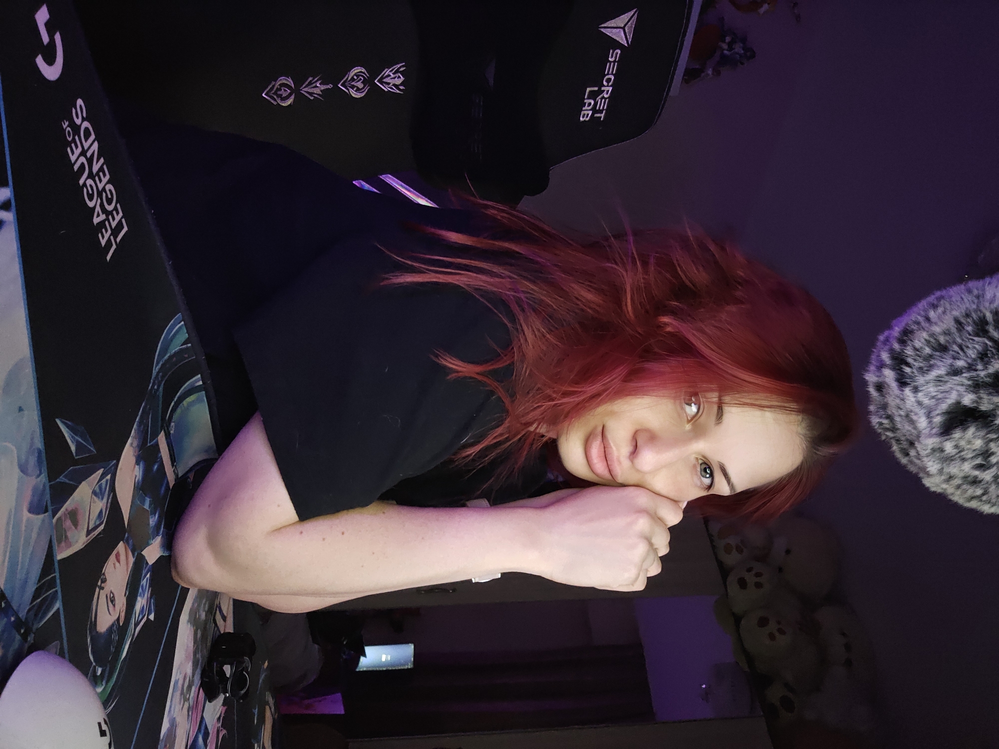 An individual with long, vibrant red hair and light-colored eyes is depicted indoors, seated at a desk in what appears to be a personal room, potentially a gaming setup, located in Burgas, Bulgaria. The individual wears a plain black t-shirt and rests their forearms on a large black desk mat. The mat features the upside-down text "LEAGUE OF LEGENDS" and a stylized illustration of a character. A white gaming mouse is partially visible on the mat. The individual's hands are clasped or positioned near the right side of their face, supporting the chin, while looking directly at the camera.
To the left, a black Secretlab gaming chair is visible, adorned with "SECRET LAB" branding and four distinct League of Legends emblems stacked vertically. The background exhibits dim, predominantly purple ambient lighting. On the right side of the frame, a shelf holds several light-colored stuffed teddy bears. Further back, a reflective surface, possibly a mirror, and a brightly illuminated vertical screen, resembling a monitor, are discernible. A large, grey, furry spherical object is partially visible hanging from the ceiling in the upper center-right portion of the image. The overall lighting suggests an evening setting or an intentionally darkened environment with accent lighting.