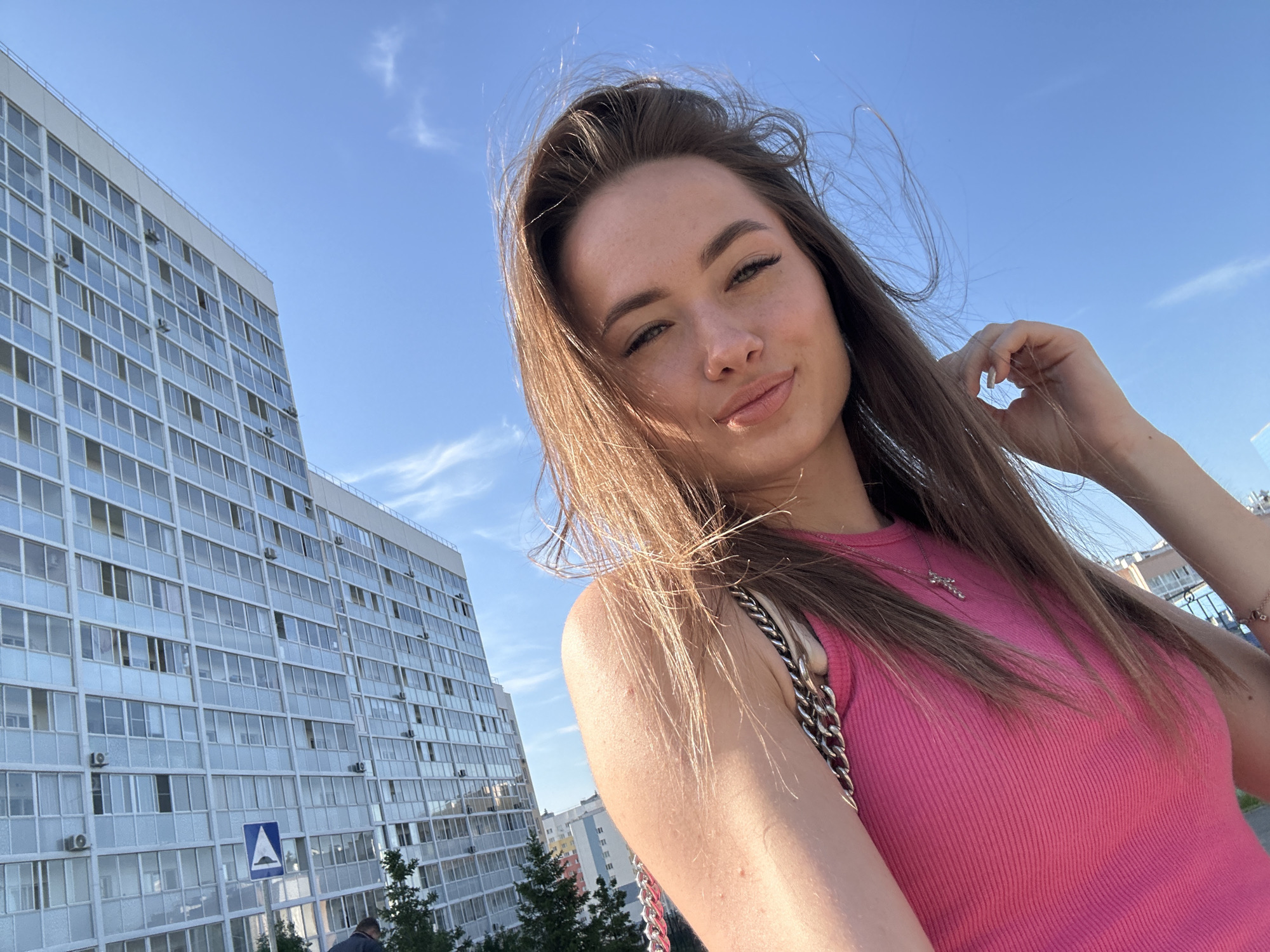 The image is a bright, sunlit selfie taken by a young woman outdoors, featuring a tall building and a clear blue sky in the background. The primary subject, a young woman, is positioned in the foreground, looking directly at the camera with a soft smile and a slight pout. Her long, dark brown hair is loose and appears to be blowing gently, with strands framing her face and her right hand holding some hair near her temple. She wears a form-fitting, sleeveless pink ribbed top and a silver-colored necklace with a small cross pendant. A silver chain strap from a bag is visible on her left shoulder. Her makeup includes defined eyebrows and eyelashes.
The setting is an urban or suburban environment on a clear, bright day. A tall, multi-story building with a light-colored facade and numerous windows occupies the left side of the background. Several air conditioning units are visible on its exterior. Below this building, a blue triangular road sign with a white upward-pointing arrow is present. Further in the background, other buildings and some green foliage are visible. The sky is a vibrant, clear blue with a few very faint wisps of clouds, suggesting either morning or afternoon. The bright sun casts a warm glow, especially on the woman's hair and skin. No specific text is clearly legible, only the symbolic road sign. The overall scene conveys a casual, lighthearted moment.