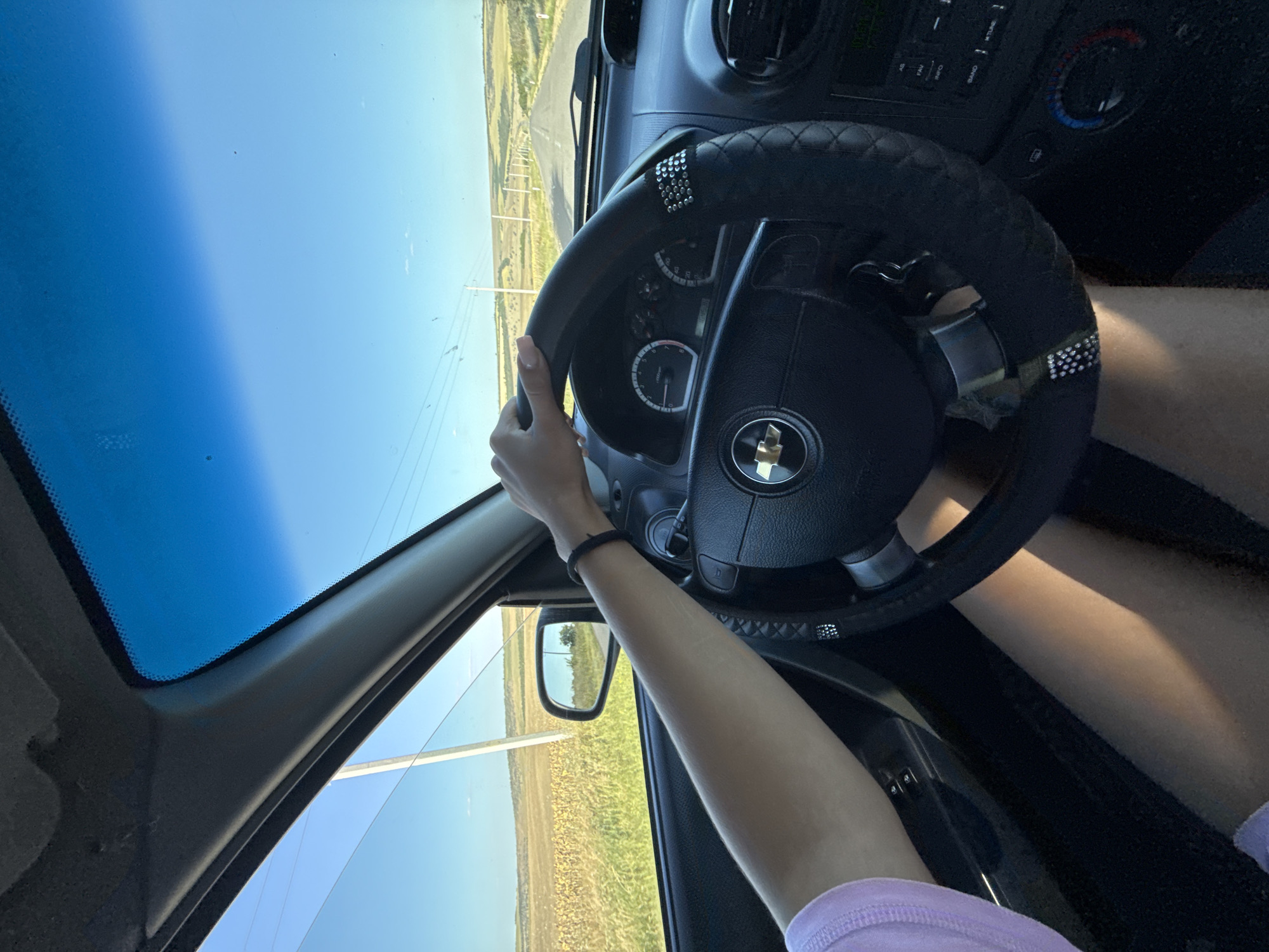 The image shows the interior of a car with a person's hands on the steering wheel. The car is driving down a rural road with fields on either side. The sky is blue and clear. There are power lines visible in the distance. The steering wheel has a black cover with decorative studs. The dashboard is visible, with gauges and a radio. The driver is wearing a light purple shirt and has bare legs. The location is identified as Cioc-Maidan, Moldova.