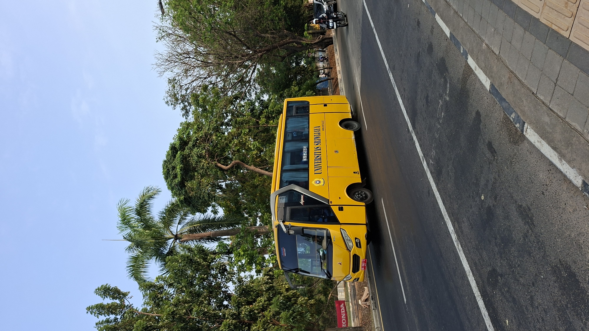 A yellow university bus with the text "UNIVERSITAS SRIWIJAYA" is driving on a paved road in Palembang, Indonesia. The bus is decorated with the university's logo and website address. The bus has a license plate that reads "BG 7493 TZ". A person is riding a motorcycle behind the bus, and a car is visible in the background. The road is lined with trees and palm trees. The sky is clear and blue. The scene appears to be during the day.