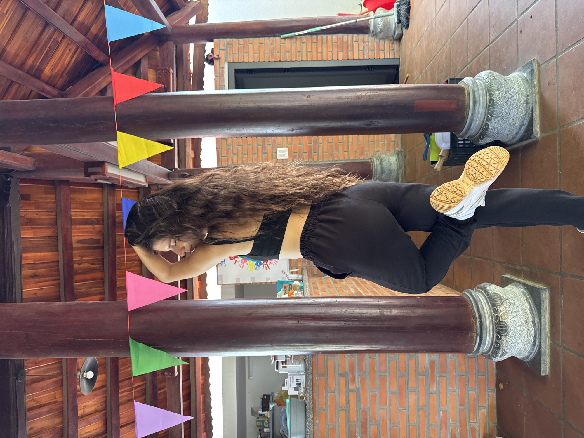 A full-body photograph captures a young woman posing casually within an open-air or semi-covered structure, characteristic of traditional Vietnamese architecture, likely a resort or communal area in Mũi Né, Vietnam. The woman, with long, wavy dark hair, stands with her back mostly to the camera, head turned to her left in profile, revealing a black sports bra and black sweatpants. Her left leg is bent with the white and beige sole of her sneaker visible, and her right hand is raised to the back of her head. She is positioned between two large, dark brown wooden pillars featuring ornate gray stone bases and black square plinths. The structure includes a wooden paneled ceiling, red tiled roof elements, and exposed red brick walls with a terracotta-colored tiled floor. Colorful pennant banners (purple, green, pink, yellow, red, blue) are strung across the pillars and ceiling beams, indicating a festive setting. To the left, a counter holds various kitchen or storage items, including a gray basin and bottles, under a black pendant light. To the right, a dark door with a gray frame, a broom, a red bucket, and a black crate are visible. A poster with colorful handprints is partially visible on the far right wall, and a small, dark object rests on a beam above the doorway.