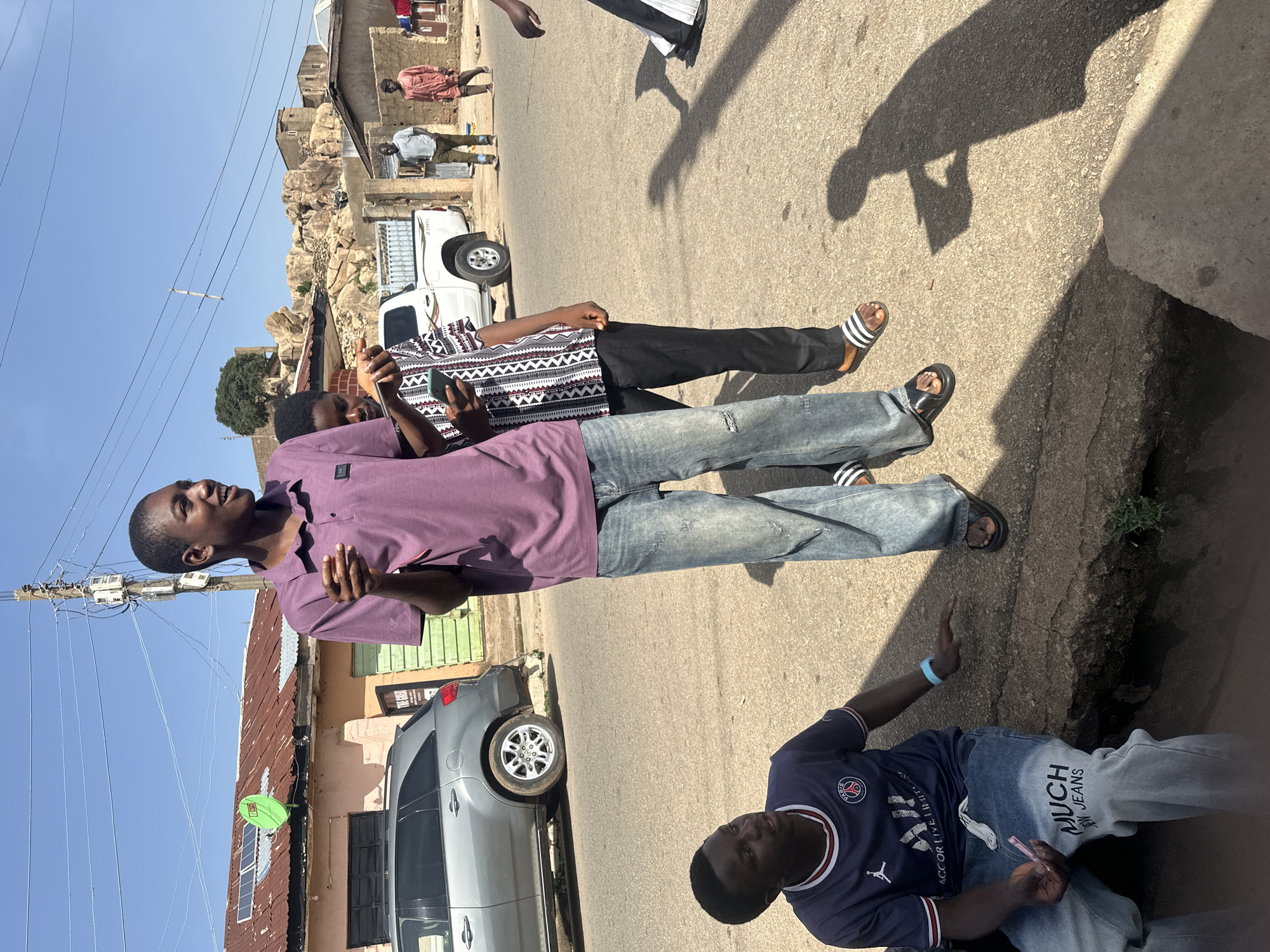The image shows a street scene in what appears to be a residential area. Three young men are standing on the side of the road. One man is wearing a purple shirt and jeans. Another is wearing a patterned black and white shirt with black pants. The third man is seated on the edge of the road wearing a blue soccer jersey and jeans. A silver SUV is parked nearby. In the background, there is a white pickup truck and buildings with rocky hills behind them. The sky is clear and blue. The location is the city of Unknown, Unknown.