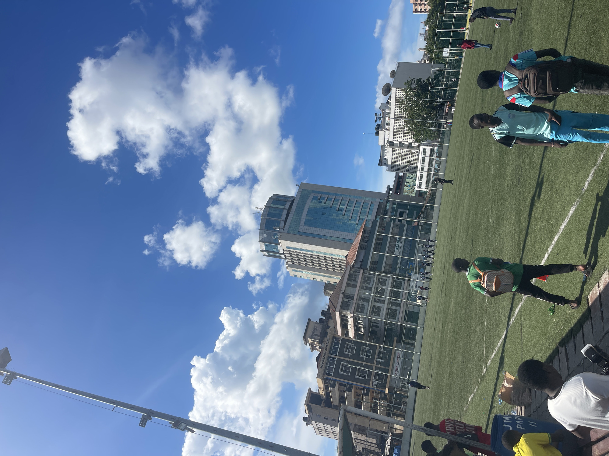 A ground-level, medium shot depicts a football (soccer) field with multiple individuals engaged in various activities against a backdrop of buildings and a blue sky with scattered clouds.
The foreground shows several people near the edge of the field. In the field, individuals are seen wearing different colored clothing, some actively playing, and others possibly observing or walking along the sidelines.
The background includes a mix of mid-rise and high-rise buildings, indicating an urban environment. A clear blue sky dominates the upper portion of the image, with scattered, fluffy white clouds.
Based on the visible signage "AZANIA" and architectural style, it is likely that this photo was taken in Dar es-Salaam, Tanzania. Buildings include Azania Bank Tower and Mlimani Building.