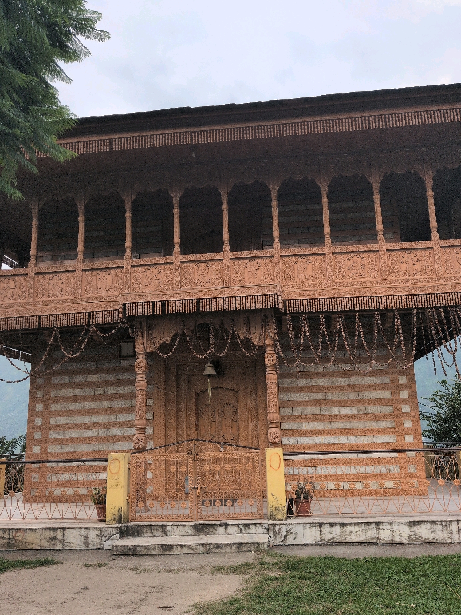 This is a full, straight-on, eye-level, medium shot of a building in Patar, India. The building is made of stone and wood, with a wooden balcony on the upper level. The balcony is decorated with intricate carvings. There is a large, ornate wooden door at the entrance, with a bell hanging above it. The building is surrounded by a garden. The sky is overcast.