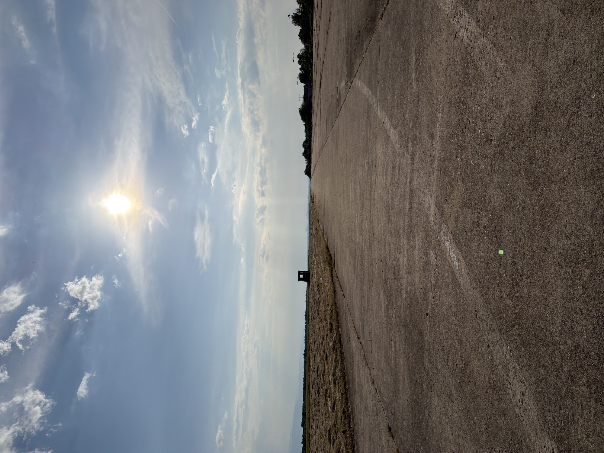 An outdoor, eye-level, wide-angle photograph depicts a long, straight, disused concrete runway or road under a bright, sunny sky in Bogdanitsa, Bulgaria. The upper half of the image is dominated by a clear blue sky with scattered white cumulus and wispy cirrus clouds. The sun is prominently featured in the upper left quadrant, casting a strong, radiant glow, with visible sun rays extending downwards. Two small, dark bird silhouettes are visible against the sky in the upper left, and a faint contrail crosses the upper right.
The horizon line is visible just below the image's vertical center. To the left, a vast field of mown, golden-brown hay or crop stubble stretches towards distant, hazy mountains. A small, dark, rectangular building, possibly a shed or watchtower, stands in silhouette on the edge of this field, roughly one-third of the way in from the left edge and just below the horizon.
The lower half of the image is taken up by the concrete surface of the runway/road, which shows signs of significant wear, cracks, and patches of sparse vegetation growing in the seams. A faint, broken white line runs down the right side of the road, curving slightly to the left as it recedes into the distance. Another, more faded white line is visible in the foreground, extending from the bottom left towards the center. A small, luminous green circular anomaly, likely a lens flare, is visible on the concrete surface in the lower right quadrant.
To the right of the road in the mid-distance, a line of overgrown trees and several tall, dark streetlights or airfield light poles are visible, extending along the perimeter of the road as it recedes into the background. No people or active vehicles are visible in the scene.