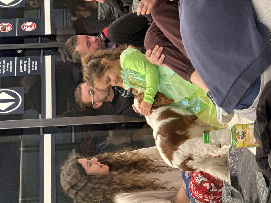 The image shows a group of individuals and a dog seated on the floor of an indoor public space, specifically identified as a light rail station in Ramat Gan, Israel. This identification is supported by safety signs in Hebrew and English affixed to a reflective glass partition in the background, which read "Do not hold on to the doors" and "Do not lean on the doors," alongside corresponding red prohibition symbols.
In the foreground, a young female child, estimated to be between 4 and 7 years old, with light brown hair pulled into a ponytail, is seated on the floor. She wears a bright green dress with a sparkly bodice. The child is actively petting a white and brown Cavalier King Charles Spaniel-type dog with both hands, displaying a broad smile. The dog is positioned between the child and an adult female seated to the left.
The adult female on the left has long, curly brown hair and wears a light-colored top. She is seated on the floor, observing the interaction between the child and the dog with a slight smile. A red and white fabric item with Mickey Mouse patterns is visible next to her.
A transparent plastic bottle of Prigat Gino brand juice with a green cap, containing a light yellow liquid, rests on the floor in front of the dog.
In the background, two adult males are seated behind the child and the dog. One male, to the left, wears glasses and a dark t-shirt. The other male, to the right, has short dark hair with grey and wears a dark top with red accents. The lower body of another individual, wearing dark brown sweatpants, is partially visible in the bottom right foreground. The floor is light-colored, and the overall environment appears clean and well-lit by ambient sources.