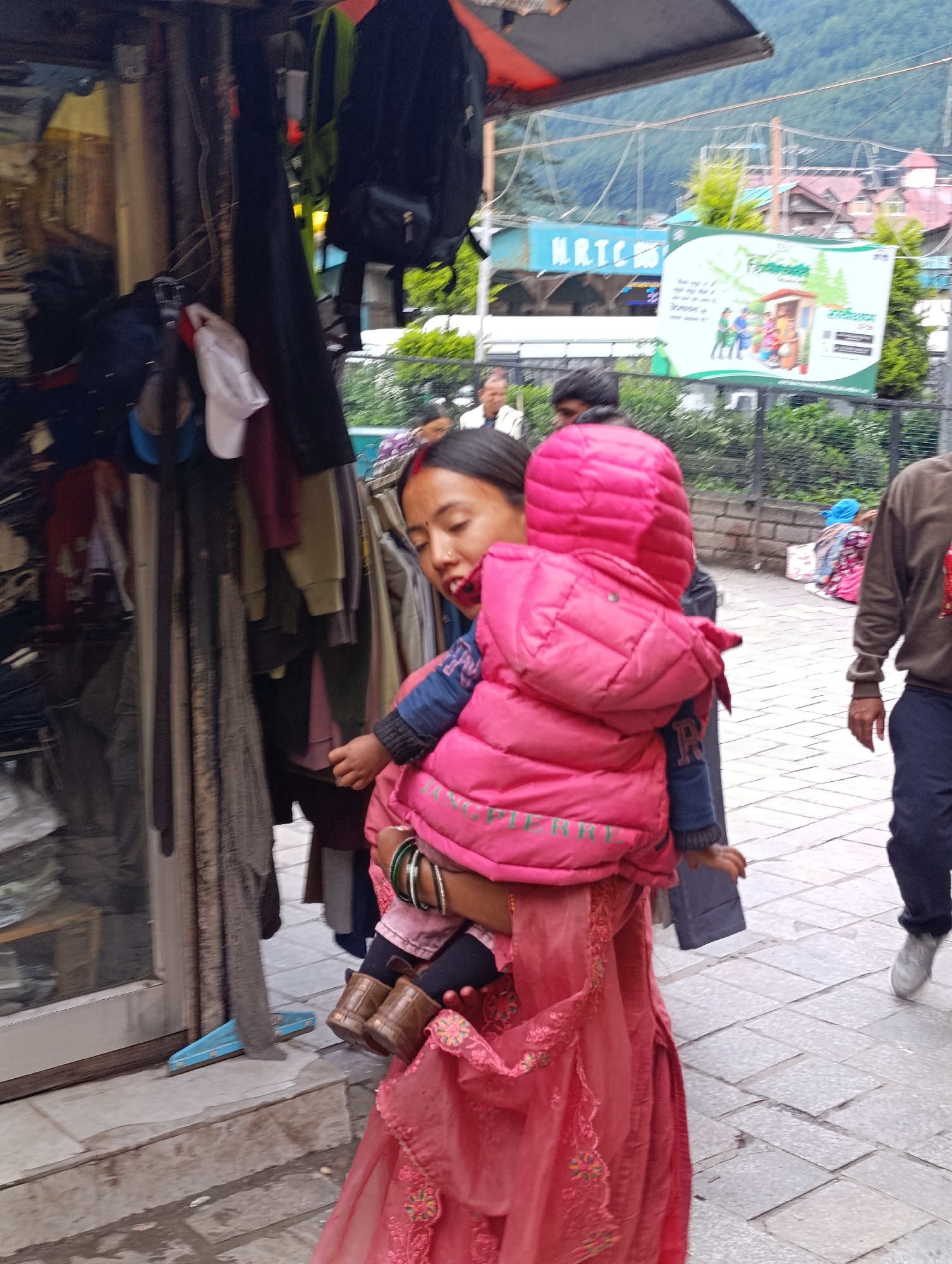 A woman with a red bindi and bangles on her wrist carries a child wrapped in a bright pink puffy jacket. The woman is wearing a pink garment with embroidered flowers. The child is wearing small brown boots. They are standing on a sidewalk in front of a shop. The shop appears to sell clothing, with racks of garments visible inside. In the background, there is a billboard with Hindi text and images, and a mountainous landscape. The scene appears to be in a busy market area in Manali, India, likely on Mall Road, a popular tourist destination.