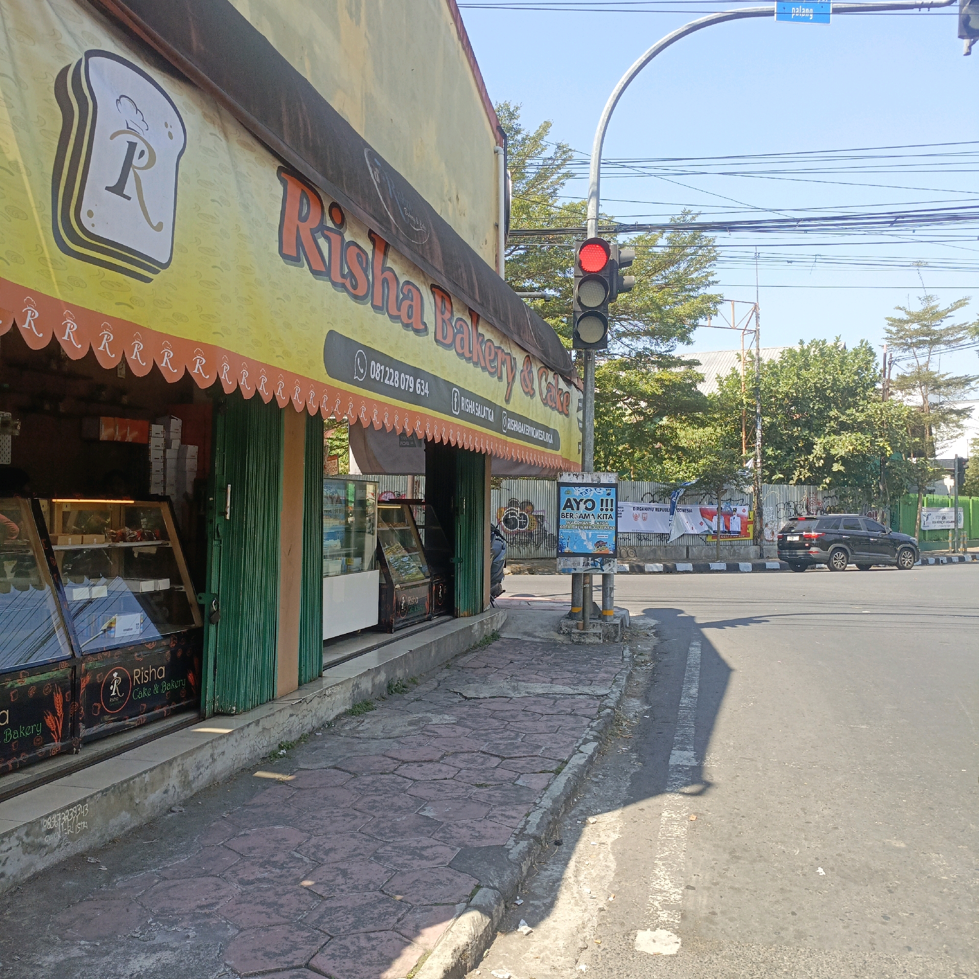 This is a street-level, eye-level, medium-shot, photograph taken on a sunny day in Salatiga, Indonesia. The image captures the exterior of "Risha Bakery & Cake," a business located on a city street. The bakery's storefront features a yellow awning with the bakery's name and logo, a loaf of bread with the letter "R" inside, and a repeating pattern of the letter "R" on a decorative orange border. A green metal roll-up door partially obscures the entrance. Display cases filled with baked goods are visible inside the bakery. To the right of the bakery, a traffic light shows a red signal. A sign on a pole next to the traffic light reads "AYO!!! BERSAMA KITA KOTA SALATIGA," which translates to "COME!!! TOGETHER WE SALATIGA CITY." A black SUV is parked on the street in the background, and banners with Indonesian government insignias are visible on a fence behind it. The sidewalk in front of the bakery is paved with hexagonal red tiles, and the street is asphalt. The environment appears to be urban, with overhead power lines and trees visible in the background.