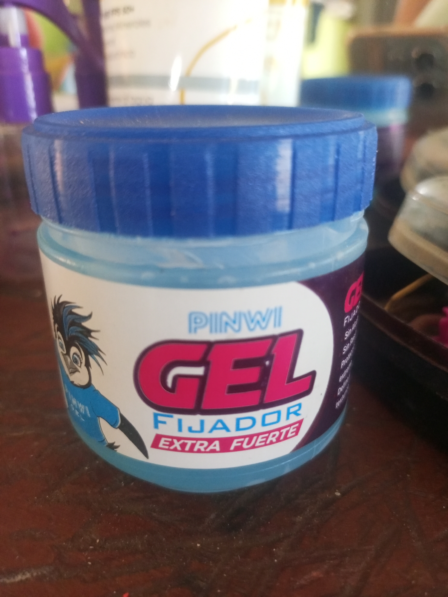 The image is a close-up, eye-level shot of a jar of "PINWI GEL FIJADOR EXTRA FUERTE" hair gel. The jar has a blue screw-on lid and a white label featuring a cartoon bird character with spiky blue hair. The label also prominently displays the brand name "PINWI" and "GEL FIJADOR EXTRA FUERTE" in large, bold lettering. The jar is placed on a dark, wooden surface that appears to be a table. The background is out of focus, showing other objects, some of which appear to be bottles or containers, also with blue caps. There are no people or discernible actions taking place in the image. The lighting is moderate, casting subtle shadows on the wooden surface. The image quality is slightly grainy. The location of La Tagua, Venezuela, is not visually represented in this image.