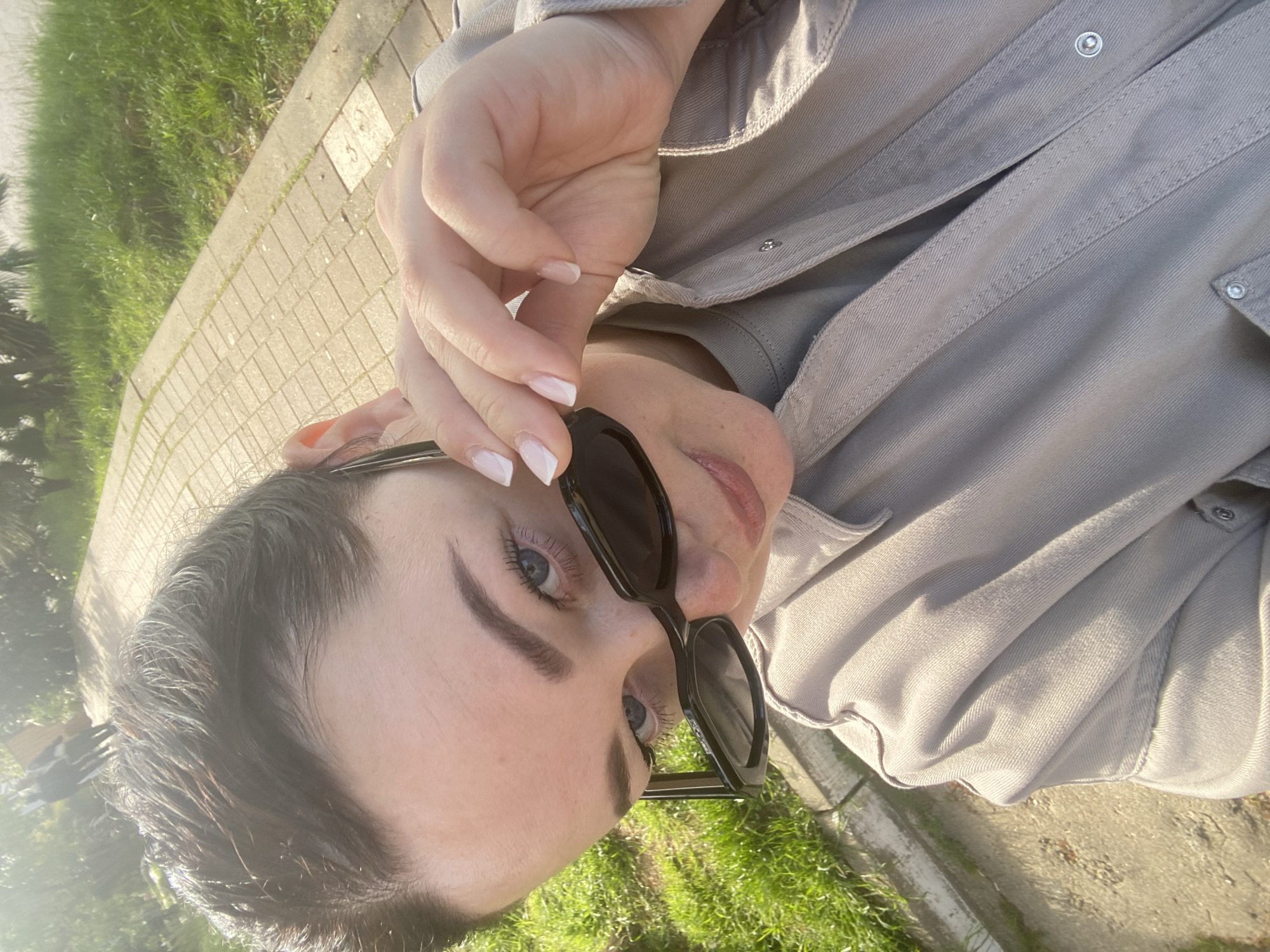 The image presents a close-up, angled outdoor portrait of a person, likely a woman, in Sochi, Russia. The subject has dark hair, striking blue eyes, and light skin, seen looking directly at the camera over a pair of black sunglasses. She holds the sunglasses slightly down with her left hand, revealing neatly manicured fingernails with white French tips.
She is wearing a light grey, long-sleeved shirt or jacket, possibly made of a sturdy fabric like denim or twill, characterized by visible stitching and snap buttons on the placket and pocket flap. The strong sunlight casts clear highlights on her hair and the fabric of her clothing, indicating a bright and sunny daytime setting.
The background shows a paved path composed of light-colored rectangular bricks, bordered by vibrant green grass. Some indistinct foliage is visible in the distance, suggesting a park, garden, or a landscaped urban area. The overall impression is a casual, direct, and slightly playful pose captured under natural daylight. There is no visible text in the image.