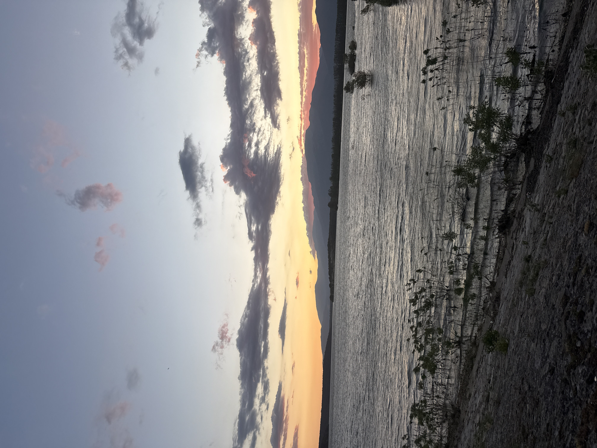 The image depicts a serene natural landscape at Koprinka, Bulgaria, likely during sunset. The sky transitions from a soft blue at the top to hues of orange and pink near the horizon, where a range of silhouetted mountains is visible. Wispy clouds, tinged with pink and gray, are scattered across the sky. A large body of water, possibly the Koprinka Reservoir, occupies the midground, its surface rippled by a gentle breeze. Along the shoreline in the foreground, sparse vegetation and rocky terrain are visible. Several small, bushy plants are emerging from the shallow water near the shore. The overall scene evokes a sense of tranquility and natural beauty. There are no people or animals present in the image.