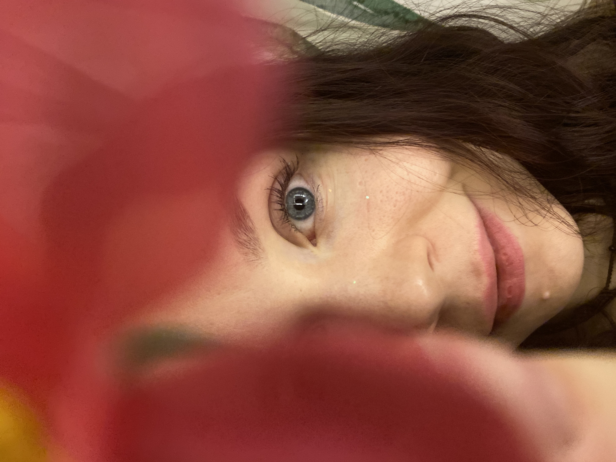 This is a close-up, rotated portrait of a person, likely female, looking directly at the camera with a subtle smile. A prominent, blurry red or pink mass occupies a significant portion of the foreground, creating a soft, obscured frame for the subject's face. The person has striking blue eyes, with the left eye (viewer's perspective) clearly in focus, showing long dark eyelashes and small specks of glitter on the skin nearby. Dark brown hair frames the upper right side of the face. Freckles are visible across the nose and cheeks. The lips are lightly colored with what appears to be pink or nude lipstick. Behind the head, blurred white and green fabric can be seen, suggesting the person is lying down on a soft surface, possibly a bed. The image conveys a casual and intimate moment. There is no visible text. The setting is indoors, in Chaikovsky, Russia.