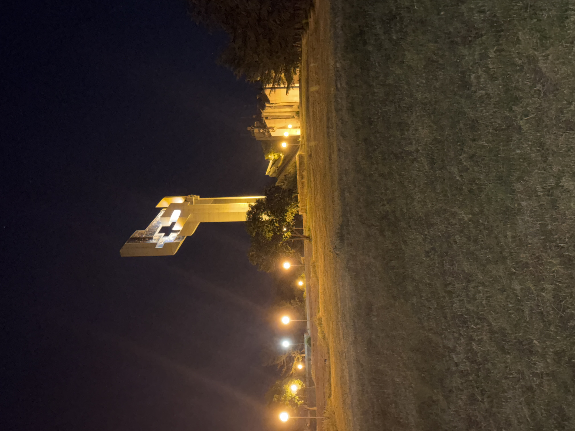 The image depicts a night scene in Stara Zagora, Bulgaria, featuring the Defenders of Stara Zagora Memorial Complex. The complex consists of a tall, rectangular structure with a stylized representation of a medieval Bulgarian flag on top. The monument is illuminated, and a row of streetlights leads toward it. A dark night sky is visible above the structure. To the right, there's a green, grassy area, possibly a park or lawn surrounding the complex. Trees are present in the scene. There are no people visible in the picture.