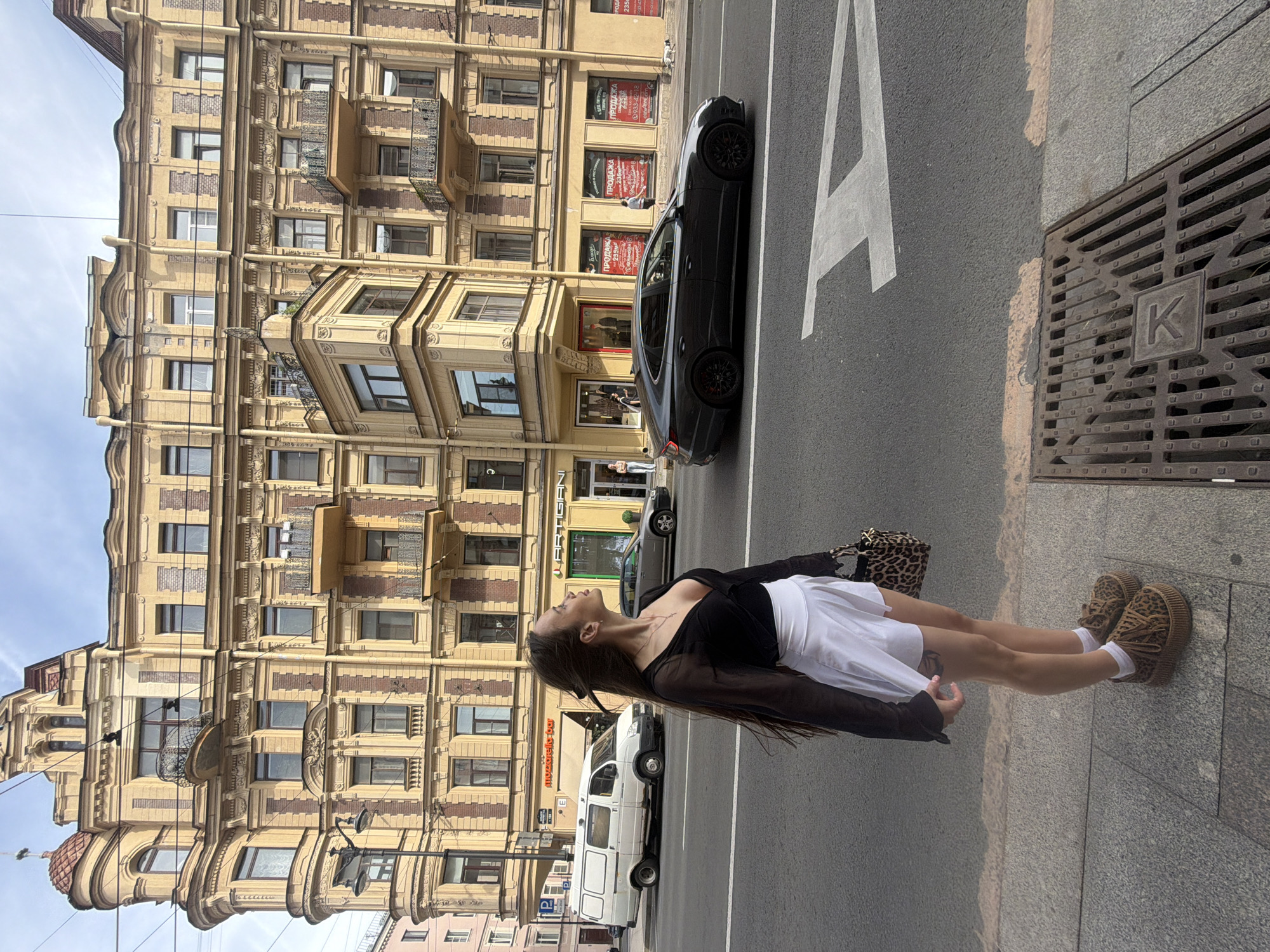 This detailed image captures a daytime urban street scene in what is identifiable as Saint Petersburg, Russia. The location is suggested by the typical architectural style of the buildings and the presence of Cyrillic script and Russian phone numbers on commercial signs.
In the immediate foreground, a young woman with long, dark brown hair stands on a concrete sidewalk, facing right and gazing upward towards the building. She is dressed in a sheer black long-sleeved top with a plunging V-neckline, a white short skirt or skort, white ankle socks, and distinctive leopard-print platform shoes. She holds a small, leopard-print handbag in her left hand. A small tattoo is visible on her upper right thigh. To her right, a rectangular metal drain cover with a raised, stylized "K" emblem is embedded in the sidewalk.
The midground features an asphalt street with a white arrow-shaped lane marking partially visible on the right. A white cargo van is parked on the left side of the road, facing left. Ahead of it, a dark grey sedan, possibly a Hyundai Sonata (not an Audi A7 as previously thought, upon closer inspection), with black multi-spoke wheels and visible red brake calipers, drives or is parked on the street, facing right. Further down the street, a lighter grey sedan is visible.
Dominating the background is a large, ornate multi-story building characterized by yellow plastered facades and red brick detailing. The building exhibits classic architectural elements, including numerous windows with decorative surrounds, several wrought-iron balconies (one notably curved at the upper-left corner, another projecting near the center-right), and various cornices. The ground floor of the building accommodates several businesses: "Mozzarella Bar" and "ARTIGIANI" are clearly visible, while other storefronts display large "ПРОДАЖА" (SALE) signs in Cyrillic, along with square footage (e.g., 255м², 235м²) and Russian telephone numbers (e.g., +7 933-42-18, +7 928-23-53). Overhead electrical wires crisscross against a sky that is partly blue with some clouds.