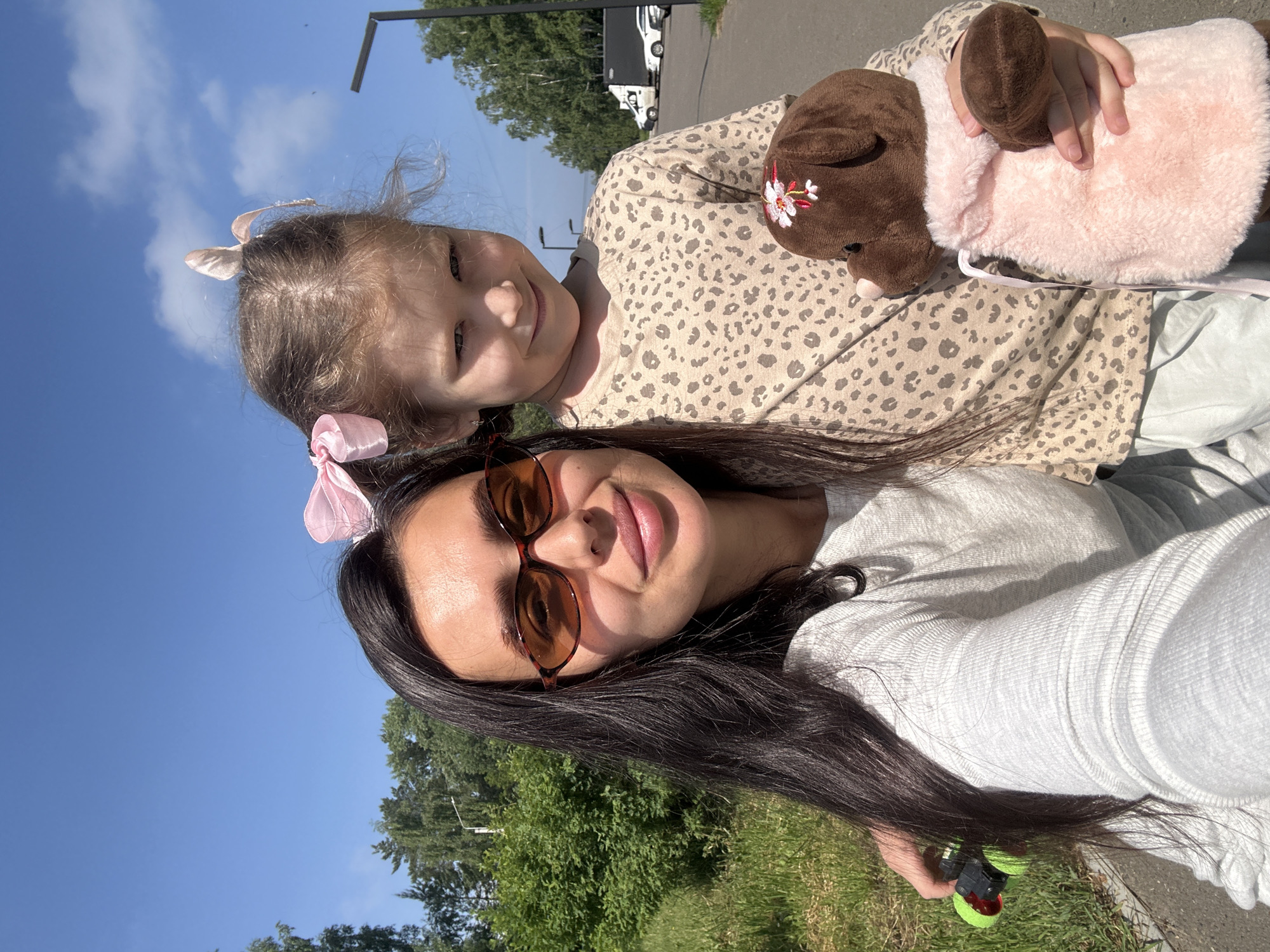 A woman and a young girl are taking a selfie outdoors on a sunny day in Chaikovsky, Russia. The woman, wearing sunglasses and a grey long-sleeved shirt, is on the left side of the frame, holding a green object with red lights. The girl, on the right, is wearing a beige leopard-print shirt and has pink bows in her hair. She is holding a brown stuffed dog with a pink furry blanket. They are standing on a paved walkway next to a grassy area with trees in the background. A lamppost and vehicles are visible in the distance. The sky is blue with some clouds.