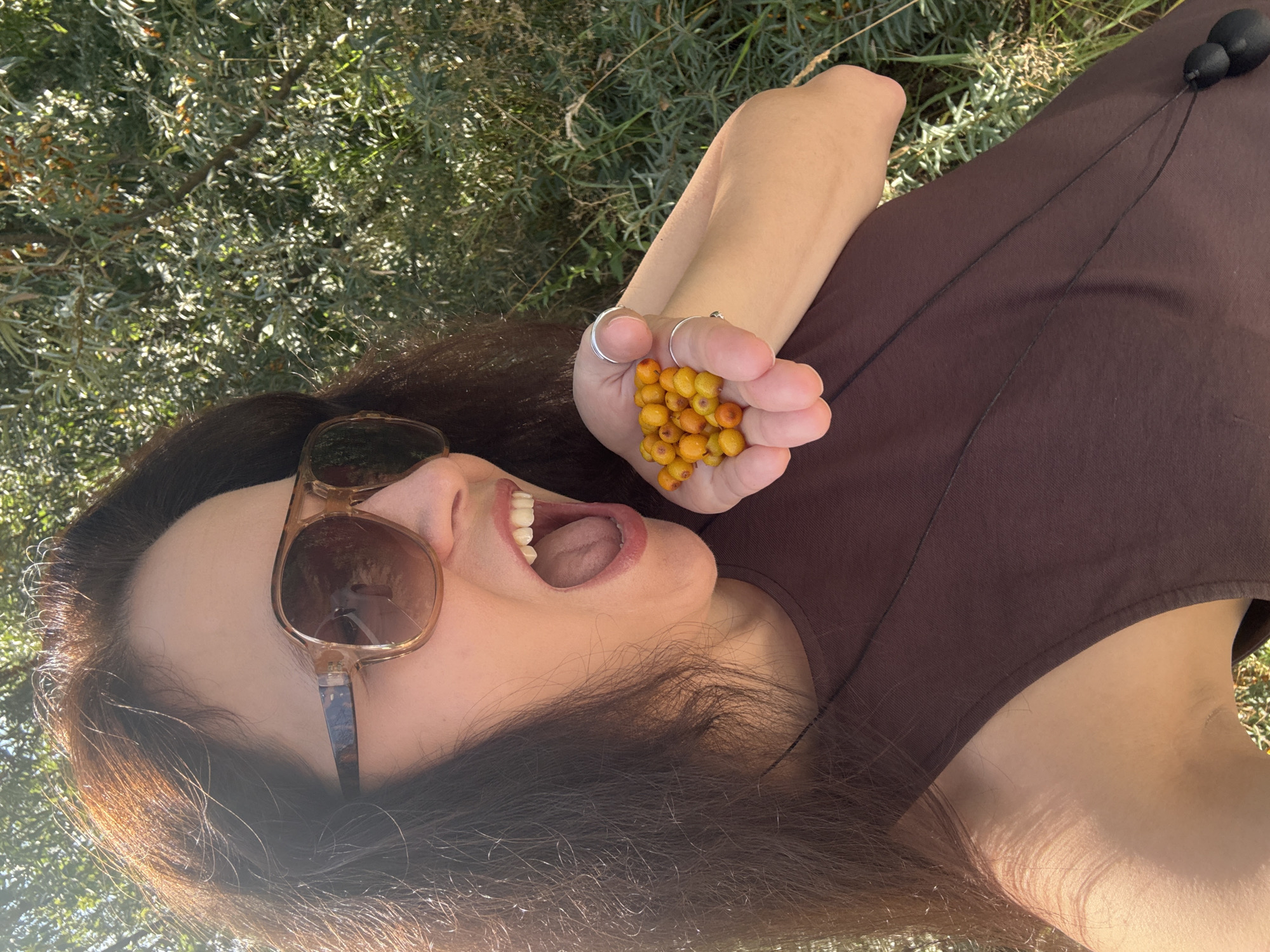 A woman with long, dark hair and large, brown sunglasses is holding a handful of small, orange berries in her right hand. She is wearing a brown sleeveless top and a necklace with two dark, round pendants. Her mouth is open, as if she is about to eat the berries. The background is filled with green foliage, suggesting an outdoor, natural setting. The image appears to be a selfie taken in daylight. There is no indication of the city of Chaikovsky, Russia within the image itself.