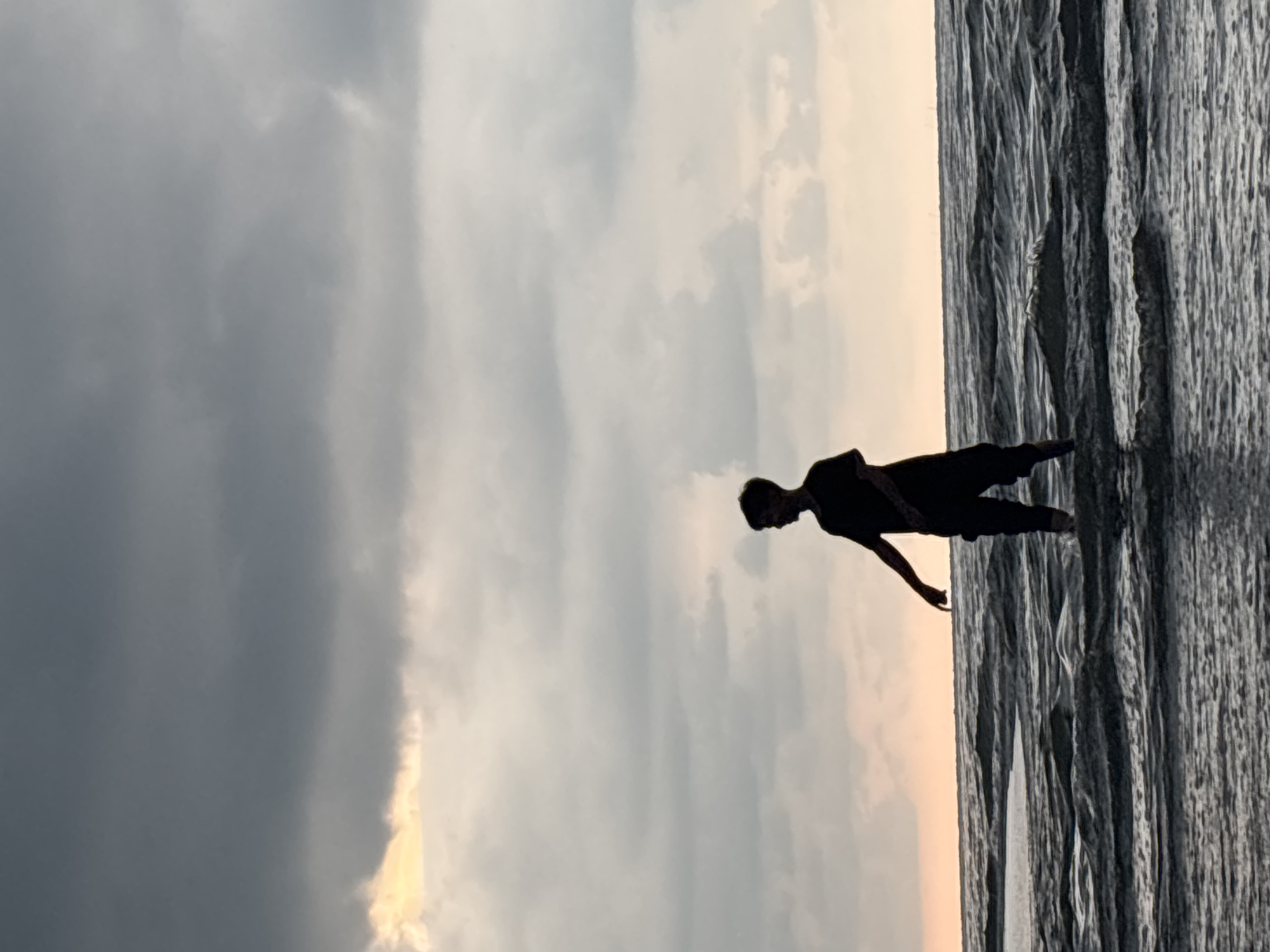 A silhouette of a person stands in shallow water at the beach, likely in Angkleng Barat, Indonesia. The sky is overcast with dark clouds above and lighter, sunlit clouds near the horizon. The water appears rippled and the image is oriented vertically.