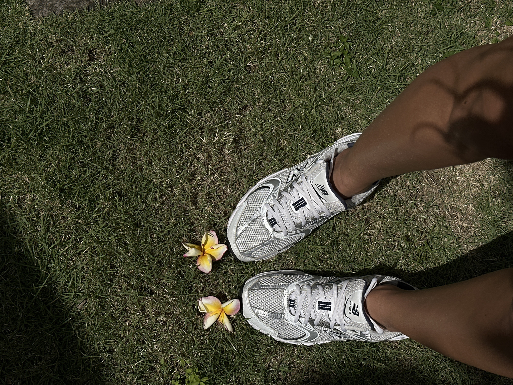 A first-person perspective shot shows a pair of white New Balance athletic shoes on a person's feet, standing on a patch of short green grass. Two plumeria flowers, one yellow and one pink and yellow, lie on the grass between the shoes. The ground is dappled with sunlight and shadow, suggesting an outdoor environment. The scene is likely located in North Kuta, Indonesia.