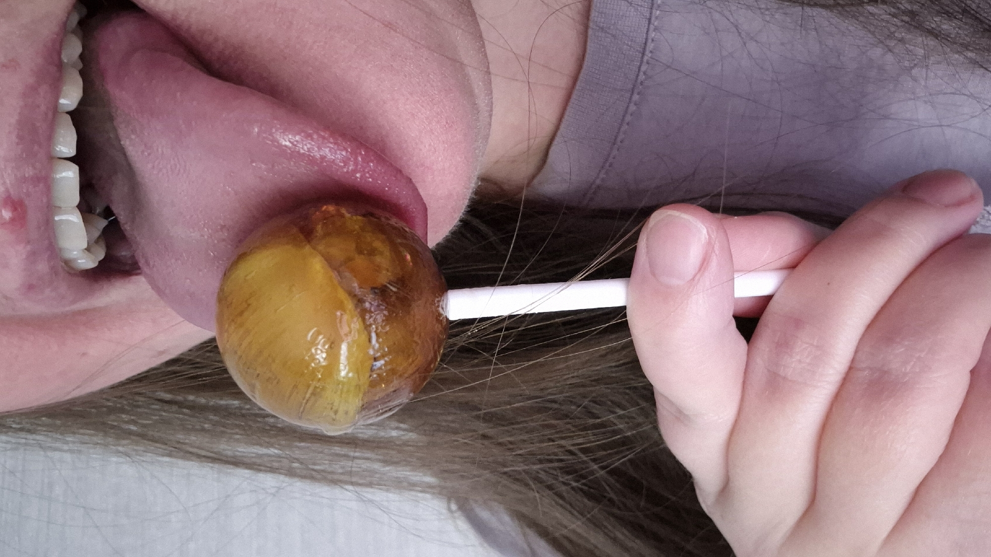 A close-up image depicts a person's mouth and hand holding a lollipop. The person's tongue is extended, licking a two-toned lollipop. The lollipop is yellow on one side and brown on the other. The person's teeth are visible. The hand holding the lollipop stick has fair skin. Hair is visible around the hand and lollipop. A portion of the person's clothing, a light purple garment, is visible above their neck.
There is no indication that this image was taken in Kemerovo, Russia.
