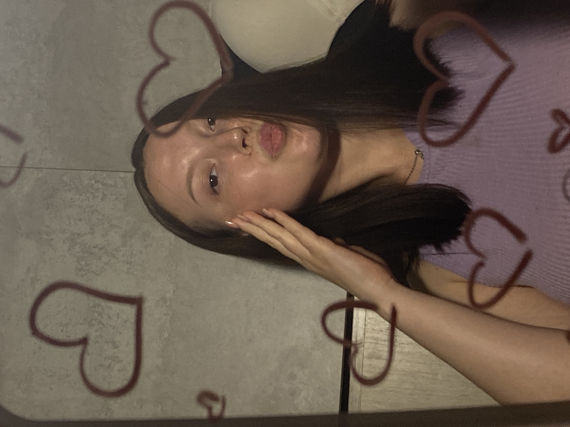 A young woman with long, dark brown hair and fair skin is posing in front of a mirror. She is wearing a lavender-colored top. Her face is close to the mirror, and she is making a kissing face. Her left hand is near her face. Dark red hearts are drawn on the surface of the mirror. The background is a textured gray wall. A partially visible beige or cream-colored object, possibly a couch or headboard, is behind the woman. The exact location in Chaikovsky, Russia cannot be determined from this image.