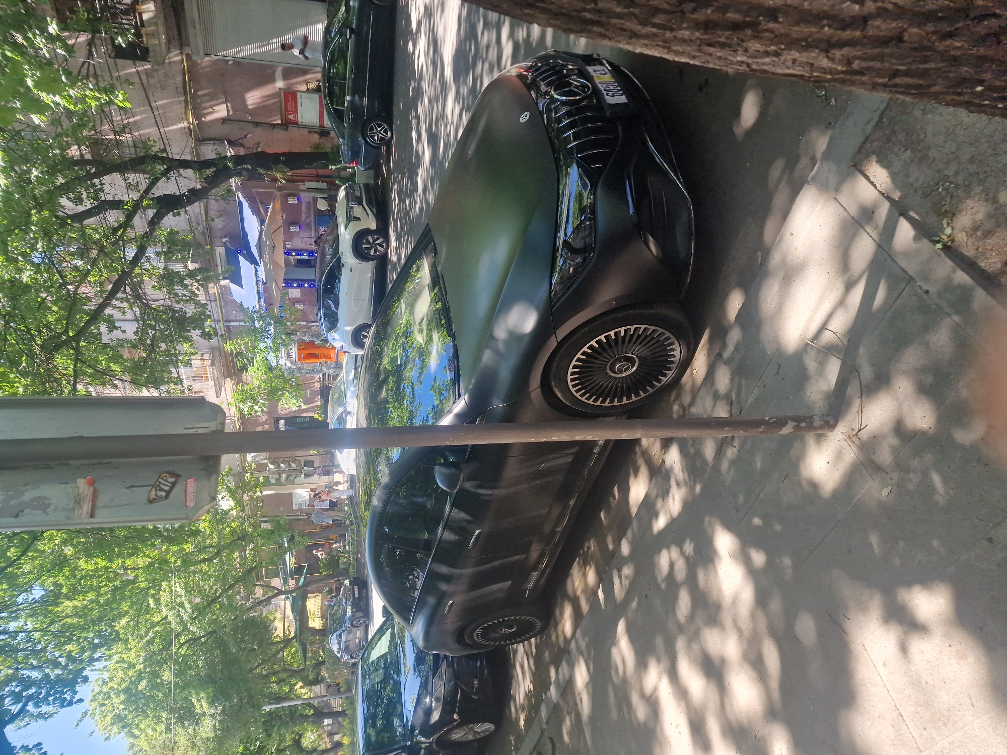 This image captures a street scene in Yerevan, Armenia, on a sunny day. The foreground is dominated by a matte black Mercedes-Benz EQS sedan, parked on the right side of the street. The car has distinctive multi-spoke wheels and a prominent grille. To the left of the EQS, another dark-colored sedan, possibly a BMW, is parked. Further down the street, a white SUV is visible, followed by a dark sedan. In the background, a multi-story building with a light-colored facade and balconies stands. Several trees with lush green foliage line the street, casting dappled shadows on the sidewalk and cars. People are visible walking on the sidewalk and near an establishment with blue awnings. A metal pole, likely a street sign or utility pole, is positioned in the center of the frame, partially obstructing the view of the cars. The overall atmosphere suggests a typical urban environment during daylight hours.