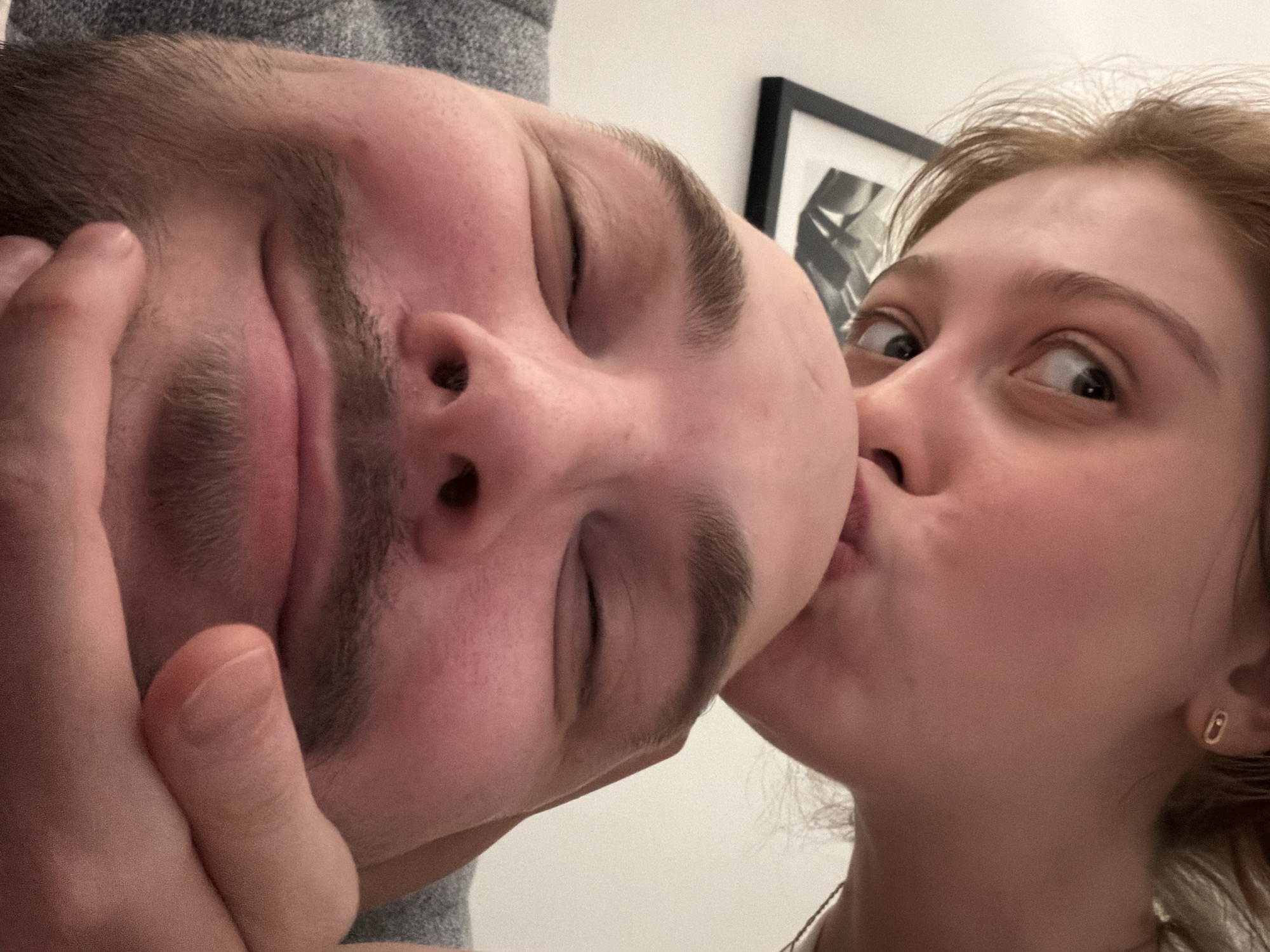 A close-up, slightly tilted selfie shows two individuals: a male and a female. The male is positioned more prominently in the foreground, his face occupying a significant portion of the image. His eyes are closed, and his hand is touching his face. He has dark hair and a mustache. The female is positioned to the right of the male, appearing to kiss his cheek. Her eyes are open, and she is looking at the camera. She has light brown hair and is wearing a small earring. In the background, slightly out of focus, is a white wall and a framed picture. There are no obvious clues to indicate that this picture was taken in Tbilisi, Georgia.