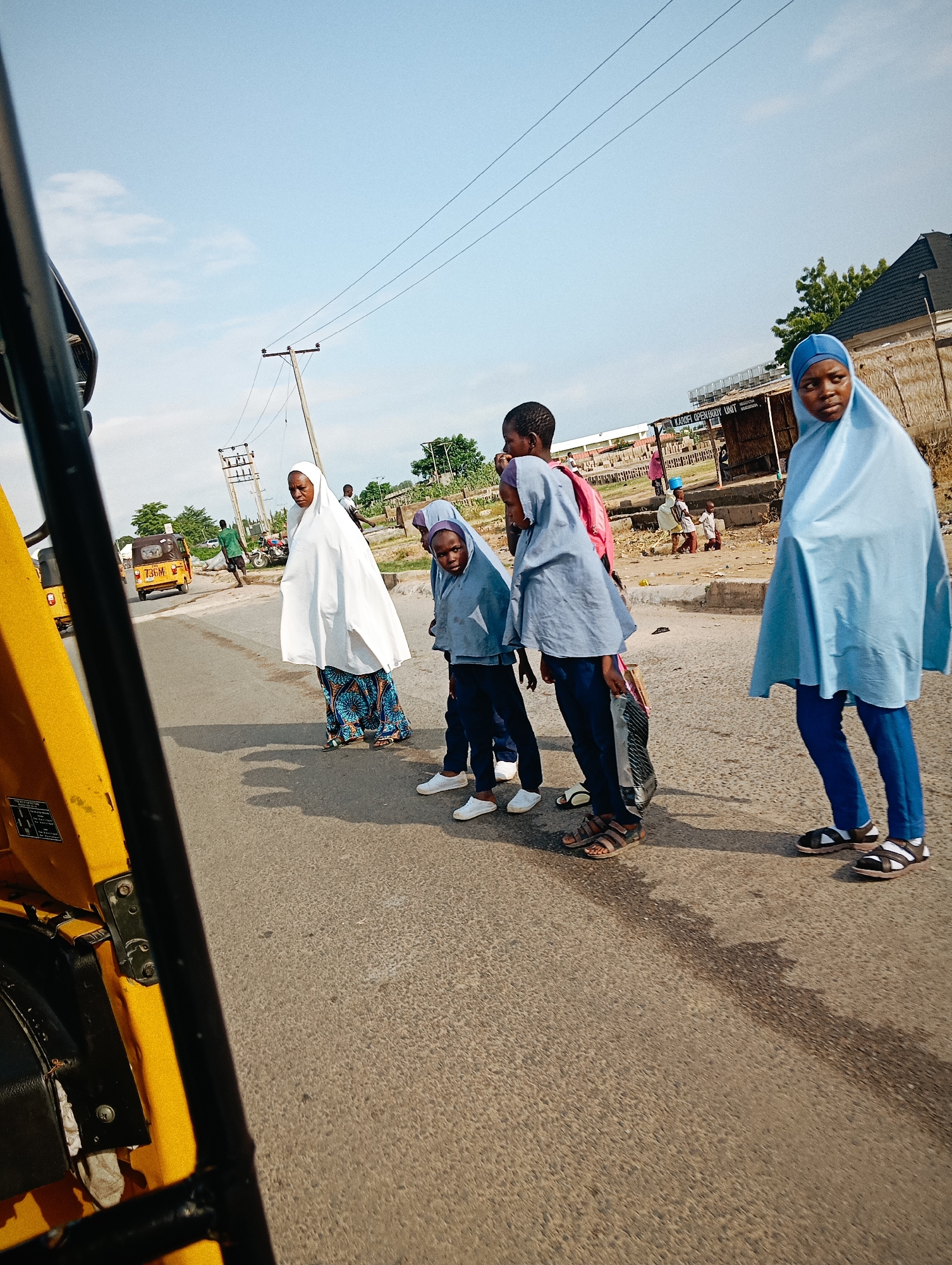 This image captures a street scene in Jalingo, Nigeria, with a prominent yellow auto-rickshaw on the left, partially obscuring the view. In the center of the road, five individuals are visible, seemingly waiting to cross or having just crossed. A woman in a white hijab and a patterned skirt stands facing left. To her right, two young girls in light blue hijabs and dark blue trousers are positioned, with one looking towards the viewer. Further to the right, another young person, also in a light blue hijab and dark blue trousers, is walking away from the viewer. The background shows a paved road, utility poles with overhead wires, and a few buildings, including one with a thatched roof. Another auto-rickshaw is seen in the distance on the road, and a person in a green shirt is walking. The sky is partly cloudy. The overall lighting suggests it is daytime.