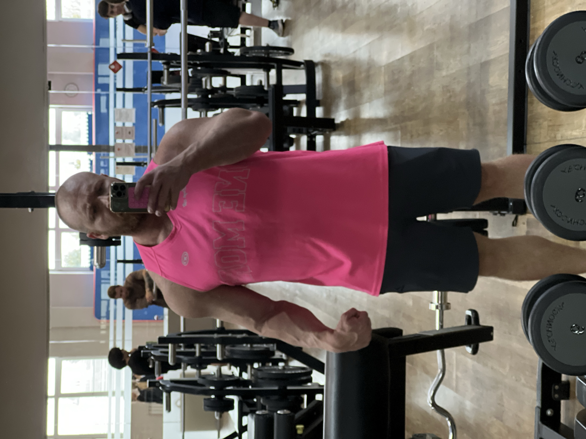 The image captures a muscular man taking a mirror selfie in an indoor gym, situated in Chaikovsky, Russia. The man is bald, has a short beard, and is dressed in a bright pink sleeveless shirt and dark shorts. He holds a silver/gray smartphone with a colorful case in his right hand, reflecting his image. His left arm is flexed, prominently displaying his bicep and visible veins.
The gym floor is covered with a light wooden or laminate material. Various pieces of exercise equipment are present, including dumbbell racks holding dark, round dumbbells. Some dumbbells bear the brand name "TECHNOGYM" and a weight indicator, with "8" being visible on one. A silver EZ curl bar is also partially visible on a rack in the background.
The large mirror reflects other elements of the gym, including glimpses of other individuals working out in the background, one in the upper right and another in the lower left of the reflection. Windows are also reflected, suggesting the photograph was taken during the daytime, combining natural light with the indoor lighting.
On the front of the man's pink shirt, stylized text is visible. In the mirror reflection, this text appears to read "MOM," but if seen directly, it would likely spell "WOW." The scene conveys a moment of fitness or a display of physical achievement.