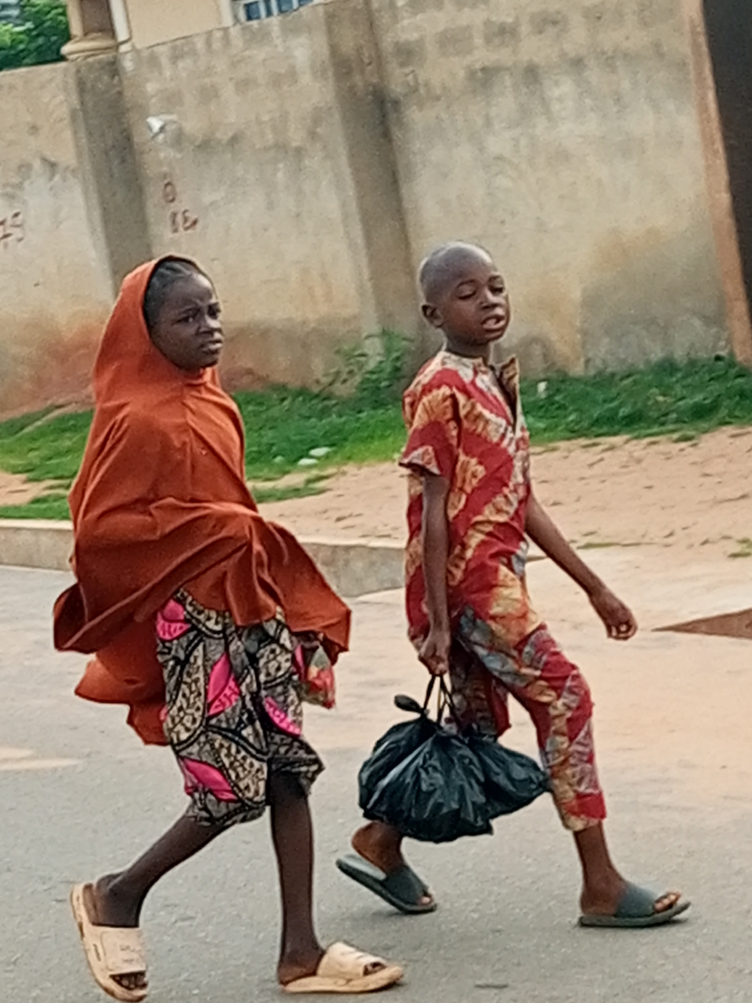 Two children are walking on a paved road in what appears to be a residential area. The child on the left is a girl wearing a rust-colored headscarf and a patterned skirt. She is looking to her left. The child on the right is a boy wearing a red and yellow patterned shirt and pants. He is carrying a black plastic bag in his right hand and is looking forward. They are walking in the same direction. In the background, there is a concrete wall with some faint markings and a grassy area. The ground appears to be dirt or sand. The overall lighting suggests it is daytime. The image quality is somewhat grainy, indicating it may have been taken with a mobile device or at a distance. The setting is consistent with a location in Tempure, Nigeria, characterized by its distinctive architecture and outdoor environment.