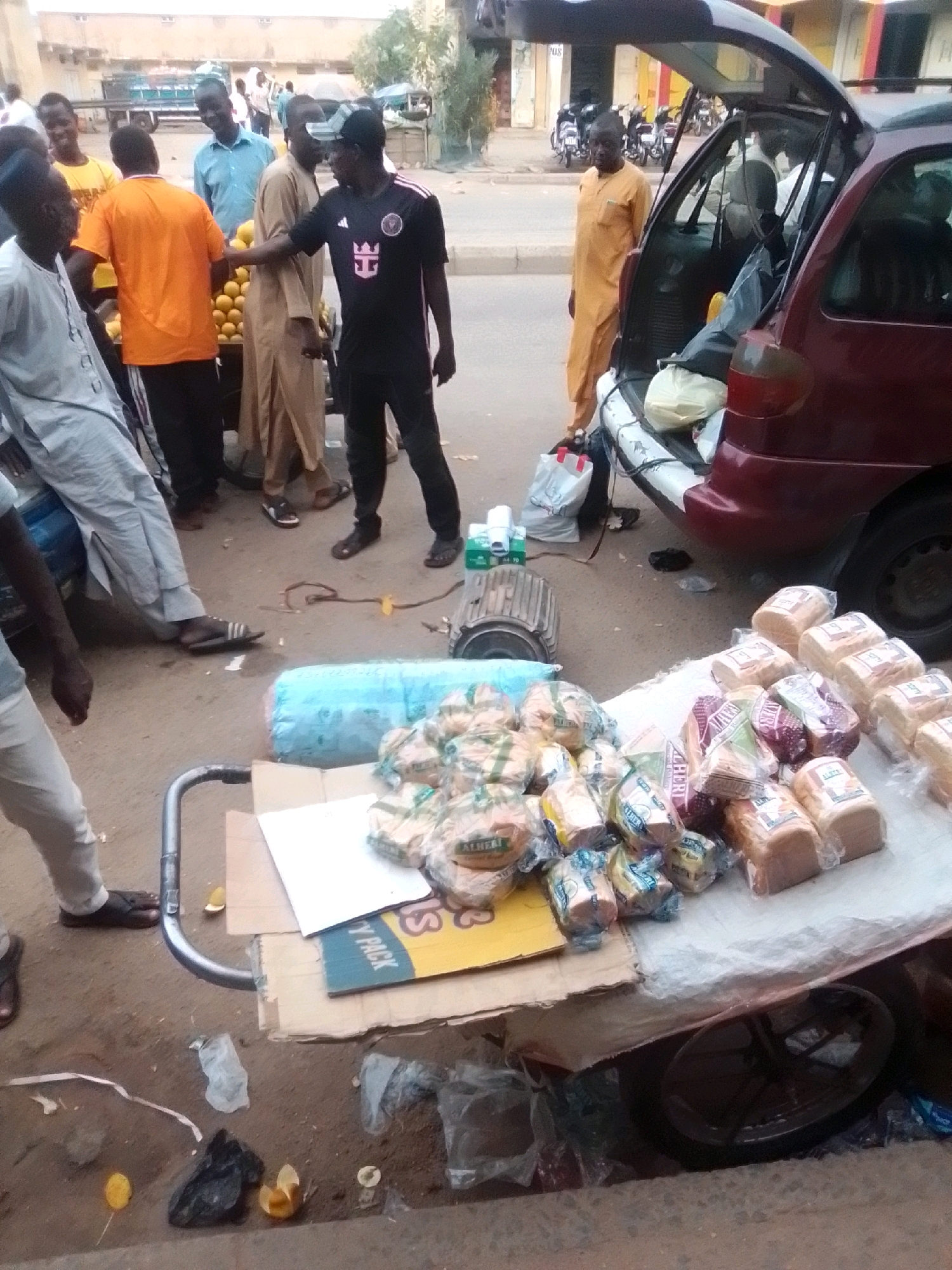 In Arawa, Nigeria, a street scene depicts a vendor selling bread from a cart next to a car with an open trunk. Several people are gathered around the cart. The vendor is wearing a black and white soccer jersey and black pants. Another person in the foreground is wearing a light gray garment. In the background, a man in an orange shirt is interacting with someone. The ground is dusty, and various items, including plastic bags and discarded food, are scattered around. The cart is laden with loaves of bread, some of which are wrapped in plastic. The open car trunk reveals more goods, possibly also food items. The overall environment suggests a bustling market or roadside selling area.