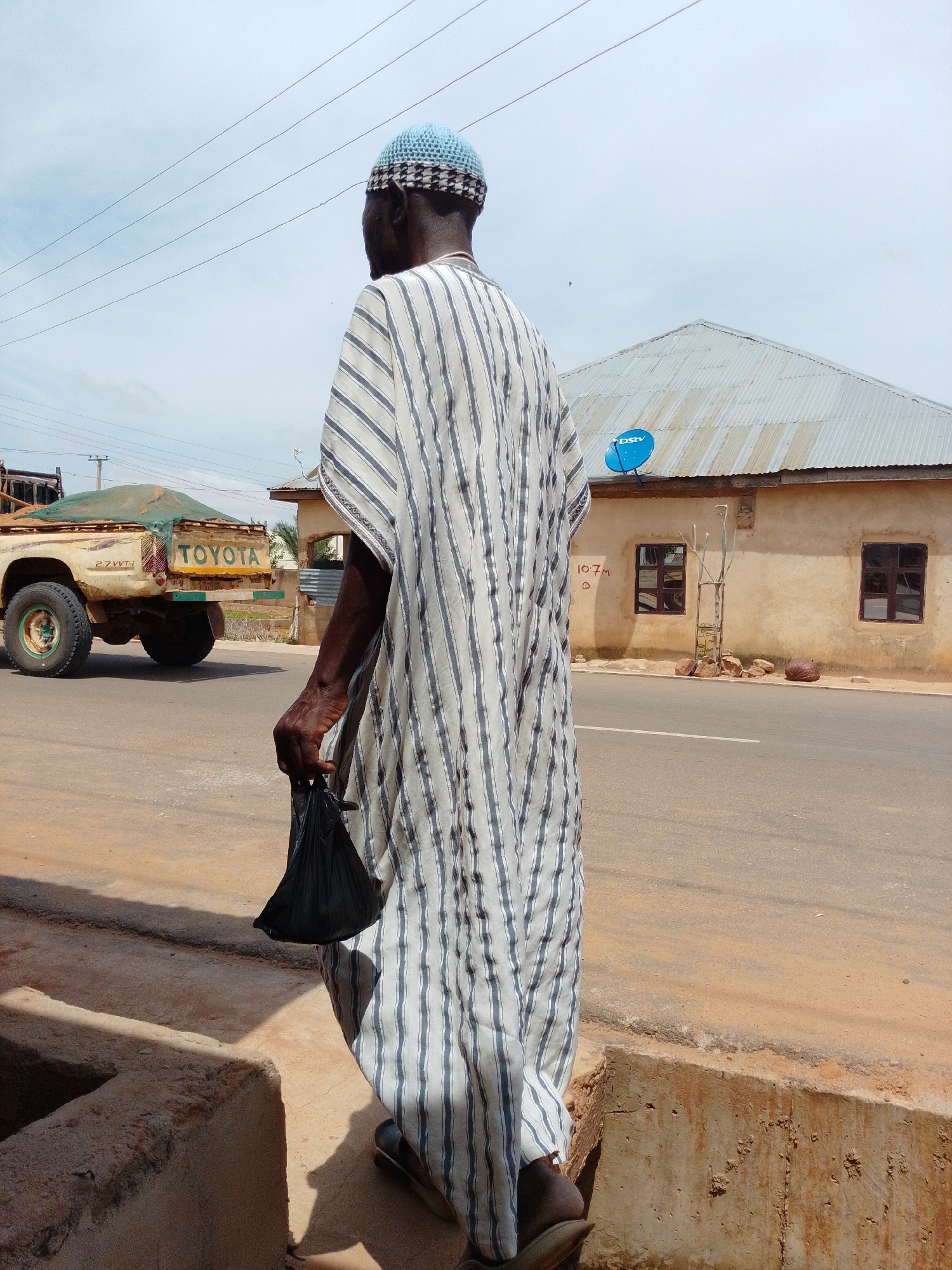 A man in a striped robe and a blue cap stands on a dirt path facing away from the camera. He is holding a black plastic bag in his right hand. Behind him is a road with a Toyota truck and a building. The building is a single-story structure with a corrugated metal roof and two windows. A blue satellite dish is mounted on the roof. The sky is overcast. The scene appears to be in a rural or semi-urban area.