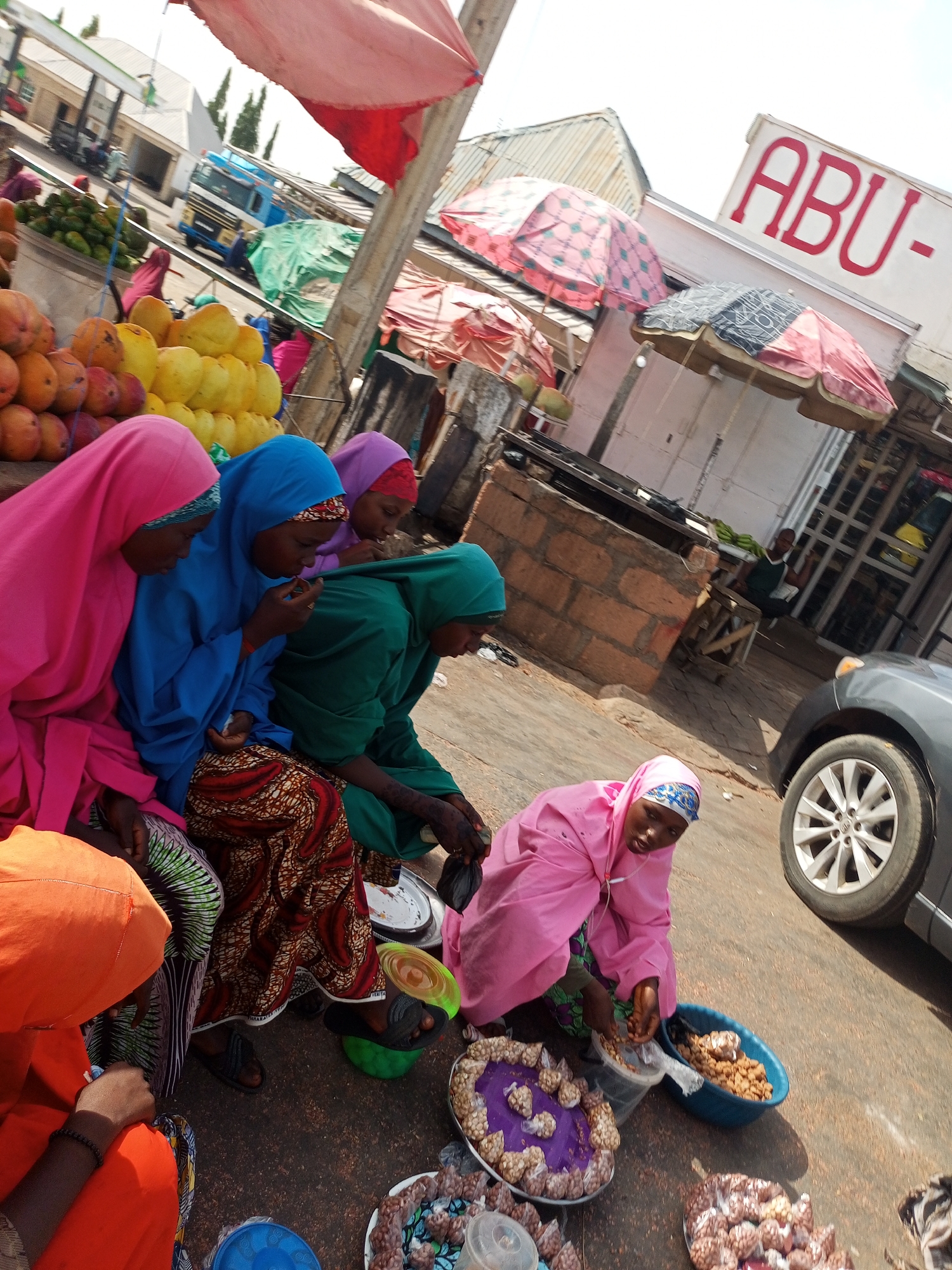 The image shows several women in hijabs of various colors, including pink, blue, green, and orange, sitting on the ground beside a road or street. They are surrounded by plates and bowls containing what appears to be food, possibly peanuts or similar snacks, packaged in small plastic bags. They appear to be preparing or selling these items.
Behind the women, there's a pile of yellow and orange fruits, possibly mangoes or similar produce. In the background, there are buildings, a truck, and umbrellas providing shade, and a car is parked on the side of the road. A sign with "ABU-" written on it is also visible. The scene suggests a roadside market or informal vending area in a urban environment in Nigeria.