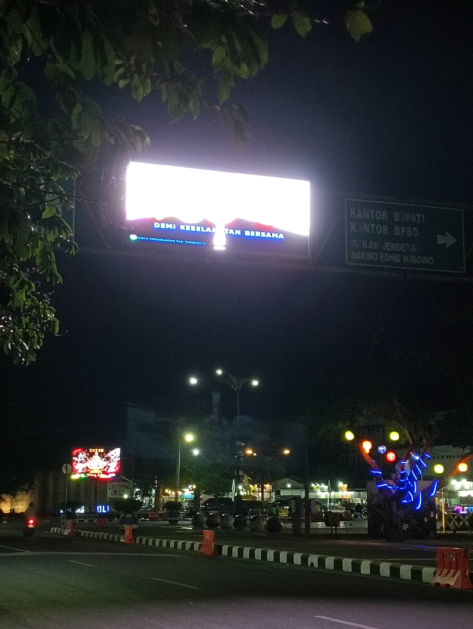 This is a nighttime, low-angle, wide shot of a street in Purworejo, Indonesia. In the foreground, a paved road with white lane markings and a striped curb is visible. Orange traffic barriers are placed along the side of the road. A lone motorcyclist is traveling away from the camera on the left side of the road. In the midground, a large, brightly lit digital billboard displays text in Indonesian that reads "DEMI KESELAMATAN TAN... BERSAMA" which translates to "FOR THE SAKE OF SAFETY TOGETHER". Below this, smaller text indicates "DINAS PERHUBUNGAN KAB. PURWOREJO", meaning "Department of Transportation Purworejo Regency". To the right of the main billboard, a green directional sign points to the right, indicating "KANTOR BUPATI", "KANTOR BPBD", and "MAKAM JENDERAL SARWO EDHIE WIBOWO", which are the Bupati's office, BPBD office, and the grave of General Sarwo Edhie Wibowo, respectively. Beyond the billboard, several buildings, streetlights, and trees decorated with colorful lights are visible. The overall impression is a busy urban intersection at night, illuminated by artificial lights.