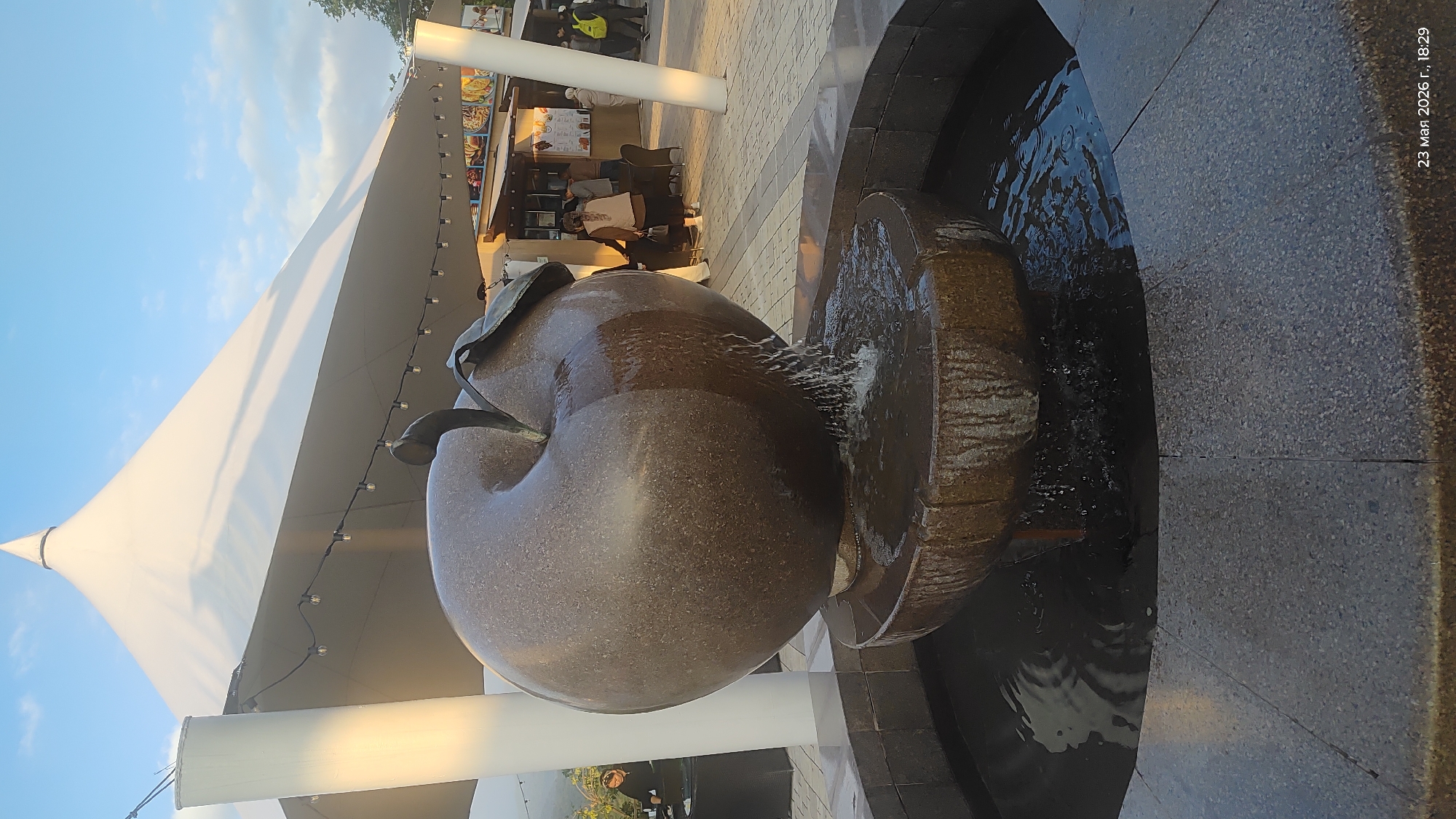 The image shows a large, apple-shaped fountain sculpture in Almaty, Kazakhstan. The sculpture is made of dark speckled stone and water is flowing from it into a surrounding basin. The basin is constructed with dark gray stone. In the background, there is a white tent structure with string lights. A food vendor stall is visible behind the fountain, with people standing and sitting nearby. The date "23 May 2026" and time "18:29" are printed on the right side of the image.