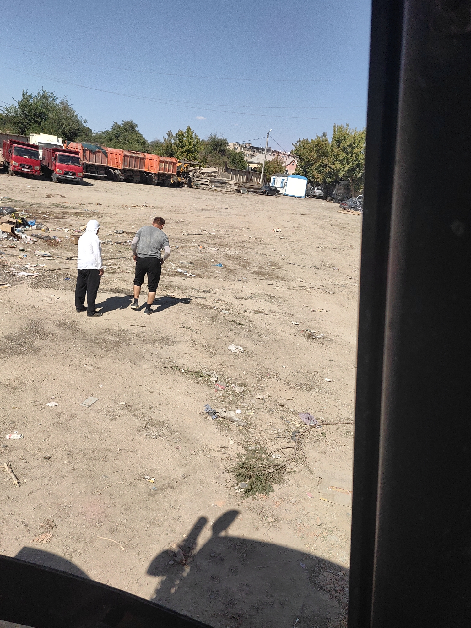 The image depicts an outdoor scene in Shymkent, Kazakhstan, likely a lot or yard. In the foreground, two individuals are present: one wearing a white hooded sweatshirt and black pants, and another in a grey t-shirt and black shorts. They are standing on a dusty, uneven ground littered with debris, including plastic bags, paper, and other miscellaneous trash.
In the middle ground, several large trucks are parked in a line. The trucks are primarily orange and red, appearing to be dump trucks or similar heavy-duty vehicles. Some of the trucks have their beds raised. Behind the trucks, there are trees and some buildings in the distance, suggesting an urban or semi-urban environment.
The overall impression is one of a somewhat neglected or working area, possibly a construction site, a storage yard, or a waste disposal area, given the presence of the trucks and the scattered debris. The sky is clear and blue, indicating a sunny day. The right side of the image is partially obscured by a dark, vertical object, possibly part of a structure or a vehicle.