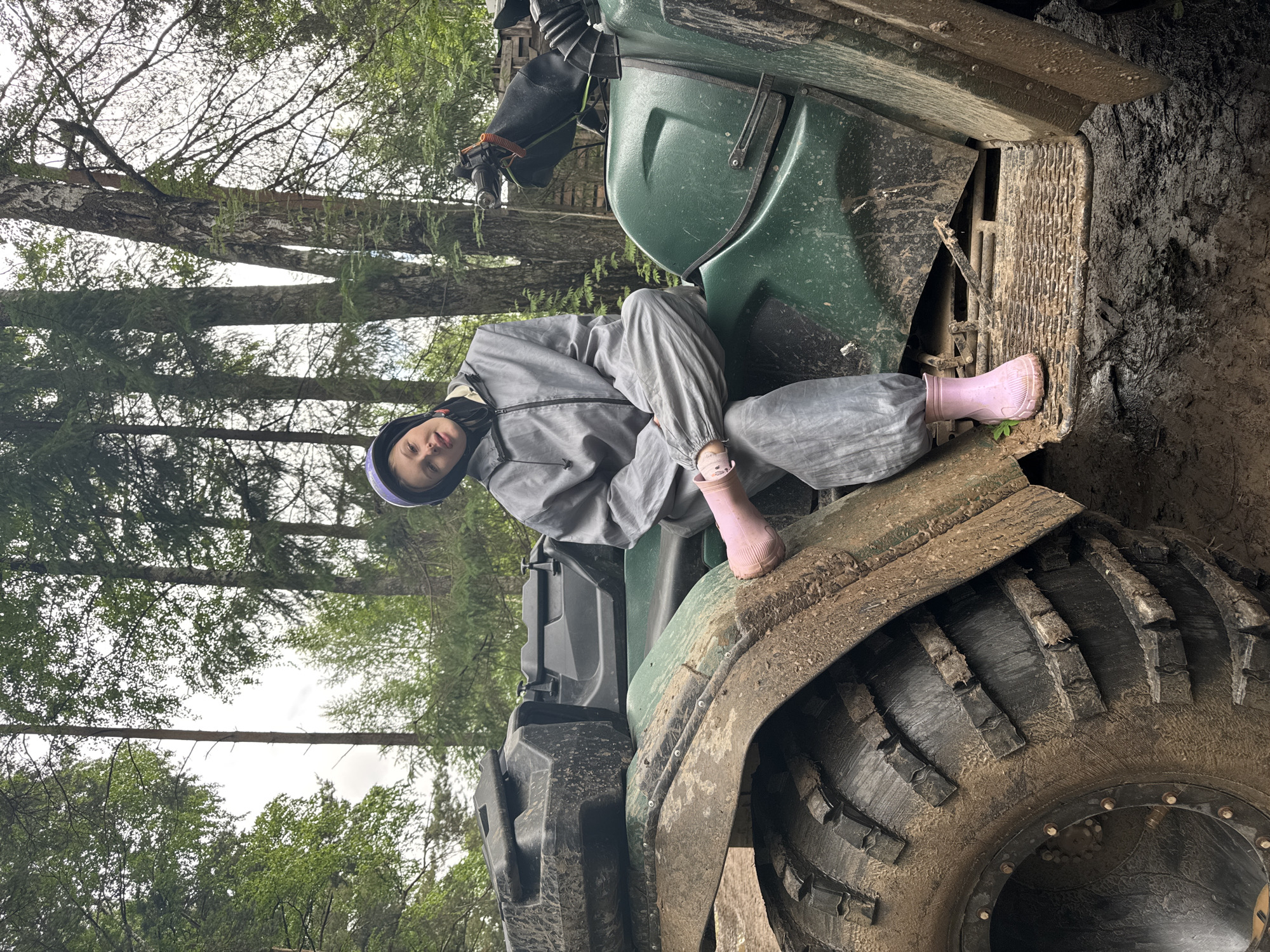 This image, captured in Perm, Russia, depicts a young female subject seated on a muddy green all-terrain vehicle (ATV) within a dense forest. The subject, with light skin and dark hair, wears a purple protective helmet, a grey full-body protective suit, and pink rubber boots. She is positioned with her legs crossed on the ATV's seat, looking directly towards the camera with a slight smile. The ATV is heavily covered in dark brown mud, particularly on its large, knobby front tire, mudguard, and footrests, indicating recent use in wet terrain. A black storage box is visible on the rear rack of the ATV. The background features numerous tall trees with green foliage, typical of a natural wooded area, under what appears to be an overcast sky. The ground surrounding the ATV is also muddy.