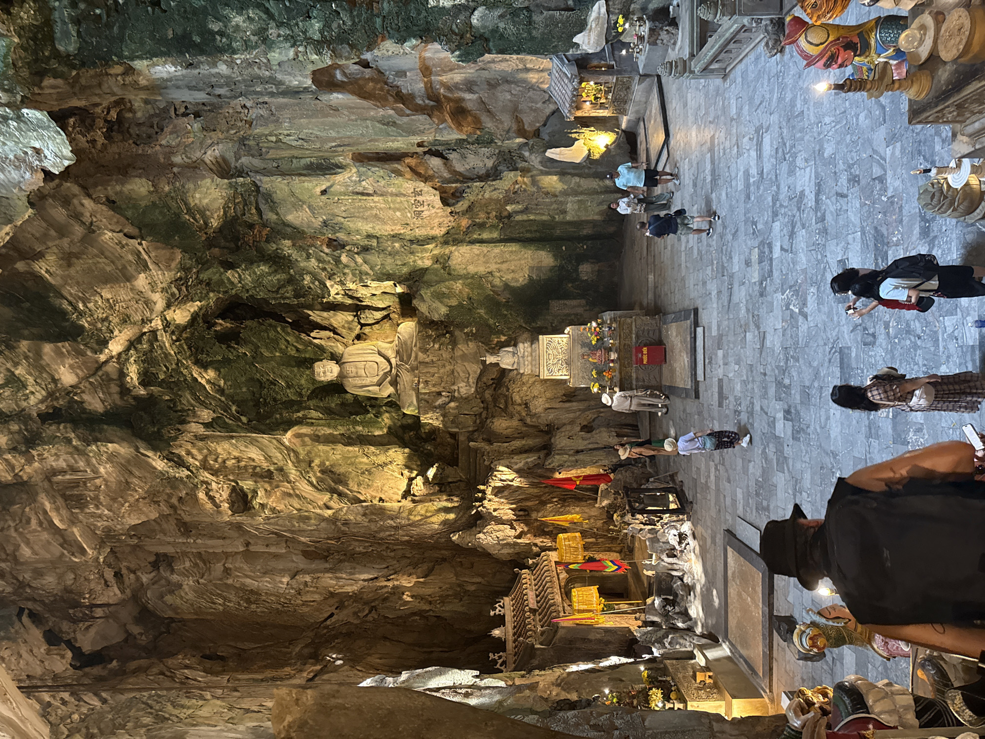 The image depicts a cave temple in Đà Nẵng, Vietnam. The cave features rock formations with a statue of a seated Buddha carved into the wall. The temple consists of an open area with a tiled floor where several people are walking, standing, and praying. Various altars and shrines are adorned with offerings such as fruit and candles. Some people are taking photos of the temple. A man in a hat is in the foreground, near several religious statues.