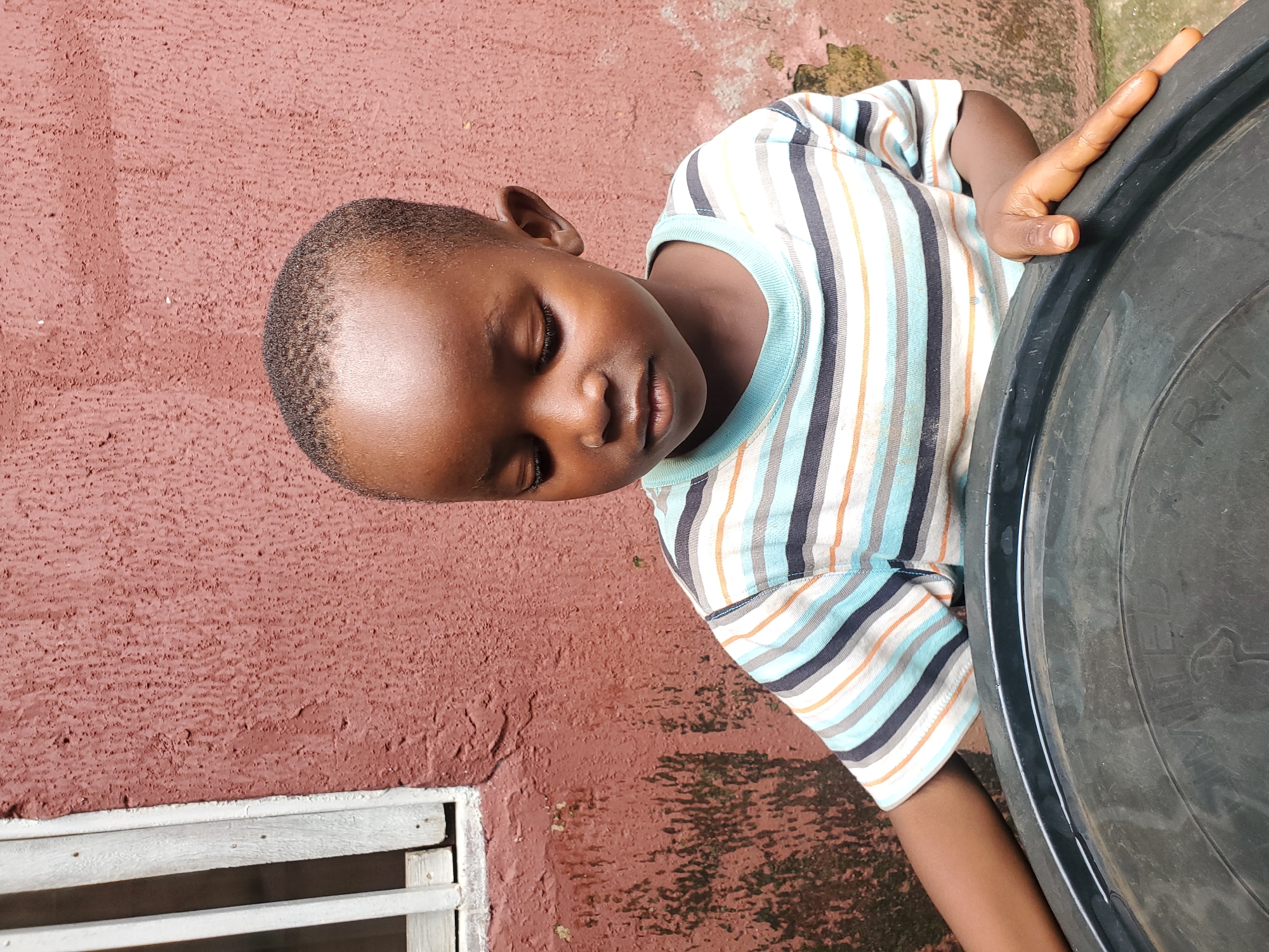 A young boy with dark skin and short hair is looking down with his eyes closed, appearing to be resting or sleeping. He is wearing a striped t-shirt with horizontal bands of white, blue, orange, and black. His right arm is resting on a large, black, circular object, possibly a container or barrel, which takes up a significant portion of the right side of the frame. The boy is positioned in front of a textured, reddish-brown wall. To the left of the wall, there is a white, slatted structure, which could be part of a window or vent. The scene appears to be outdoors, given the natural lighting and the texture of the wall which suggests an exterior surface. The overall impression is one of a quiet, possibly warm moment, as the boy seems relaxed. The image quality is consistent with a photograph taken with a mobile device. There is some faint text visible on the black object, appearing to be embossed letters, but it is not clearly legible. The context of Jos, Nigeria, places this scene within an urban or semi-urban environment in West Africa.