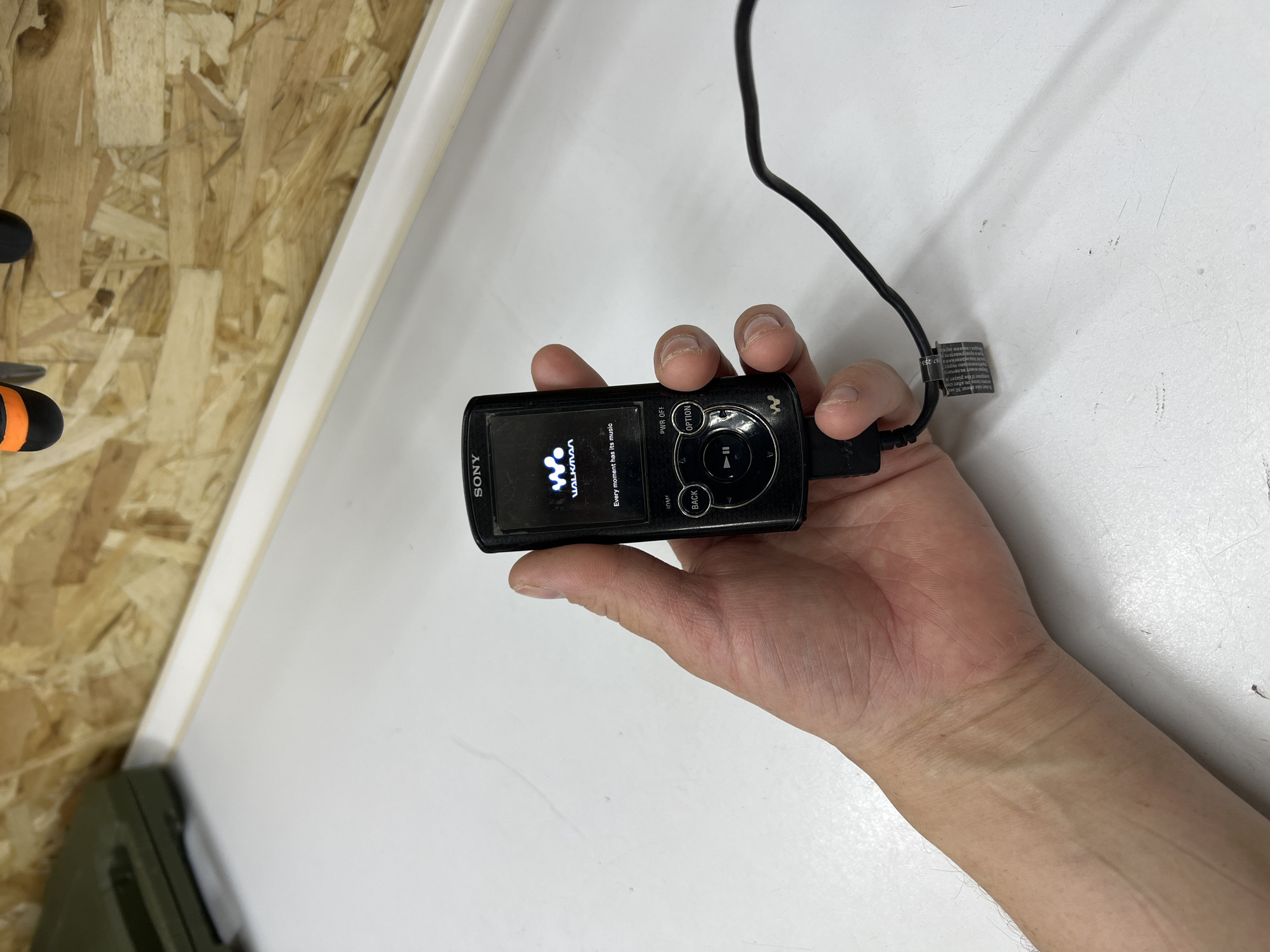 A person's left hand is holding a black Sony Walkman MP3 player. The screen of the Walkman displays a "W. WALKMAN" logo and the slogan "Every moment has its music." The device is connected to a black cable. The background shows a white surface, possibly a table, and a wooden wall. There is also a green case visible on the left side of the image. The scene appears to be indoors, and there is no indication of the location within the city of Veteran-2, Russia.