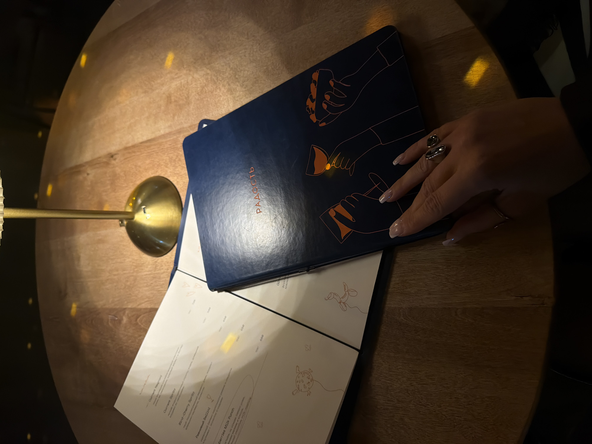 The image depicts a menu from a restaurant in Krasnoyarsk, Russia. The menu, with a navy blue cover featuring orange hand illustrations, is lying open on a wooden table. A person's hand, adorned with two large rings, rests on the menu. A lamp with a golden base is also visible on the table, casting a warm light. The open menu pages reveal various drink options, including "Current Negrond," "Current Spritz," "Bird-Cherry Spritz," "Ревнивый Bellini," and "aberries Milk Punch," each listed with its price in Russian rubles. The word "РАДОСТЬ" (Radost', meaning "Joy" in Russian) is printed in Cyrillic on the menu cover.