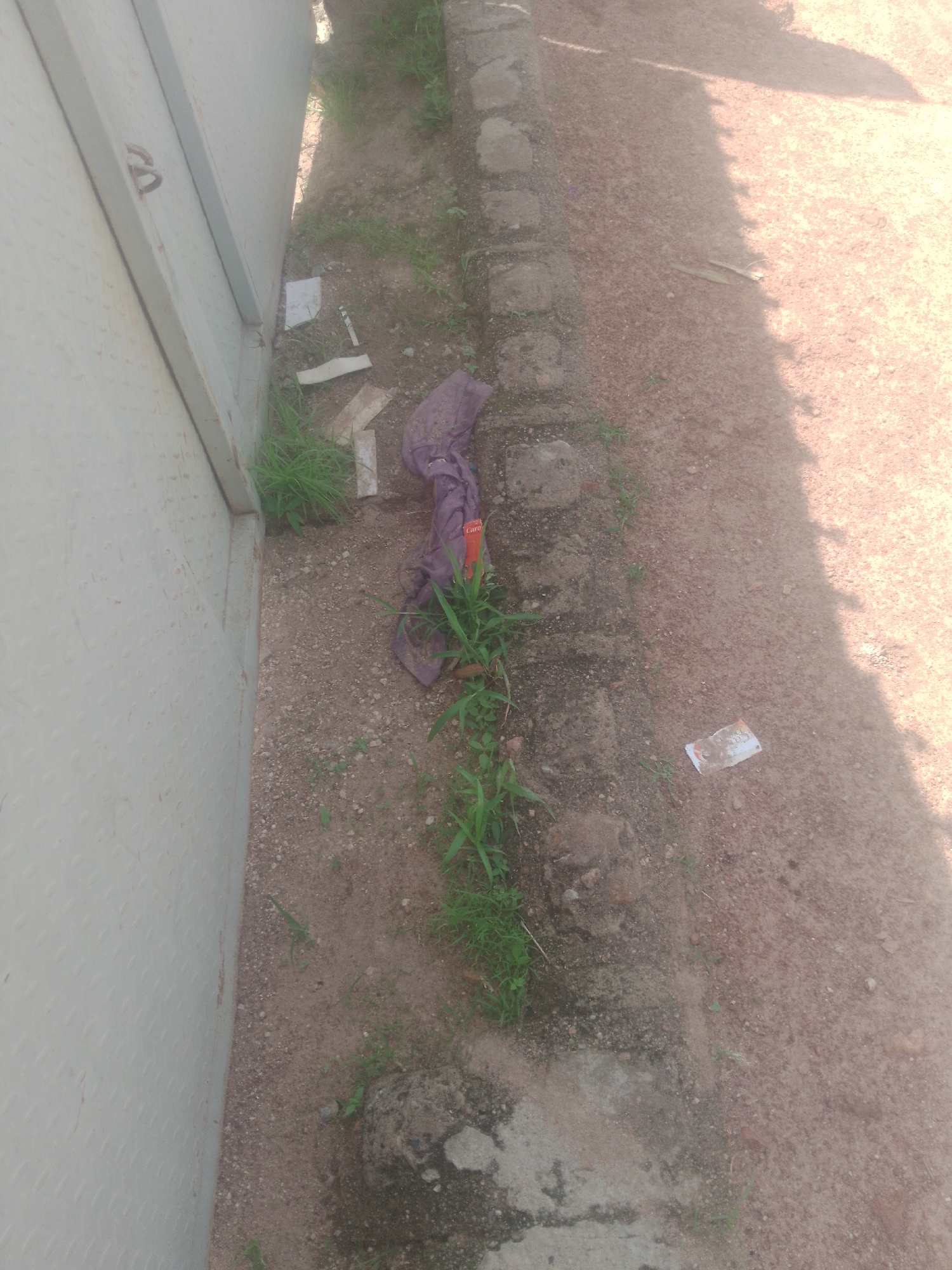 In the city of Jauro Yinu, Nigeria, a side view of a textured light-colored metallic fence is visible on the left. Adjacent to the fence, a strip of ground is lined with rough, grey concrete blocks. Beyond the concrete, the ground is a mixture of sandy soil and small stones, with sparse patches of green grass growing in places. A crumpled piece of dark purple fabric, possibly clothing, lies on the ground near the concrete border, with a small orange object and green grass intertwined with it. Further into the sandy area, a few scattered pieces of paper or thin plastic are visible, one white piece with faint writing. A distinct shadow falls across the sandy ground from the right.