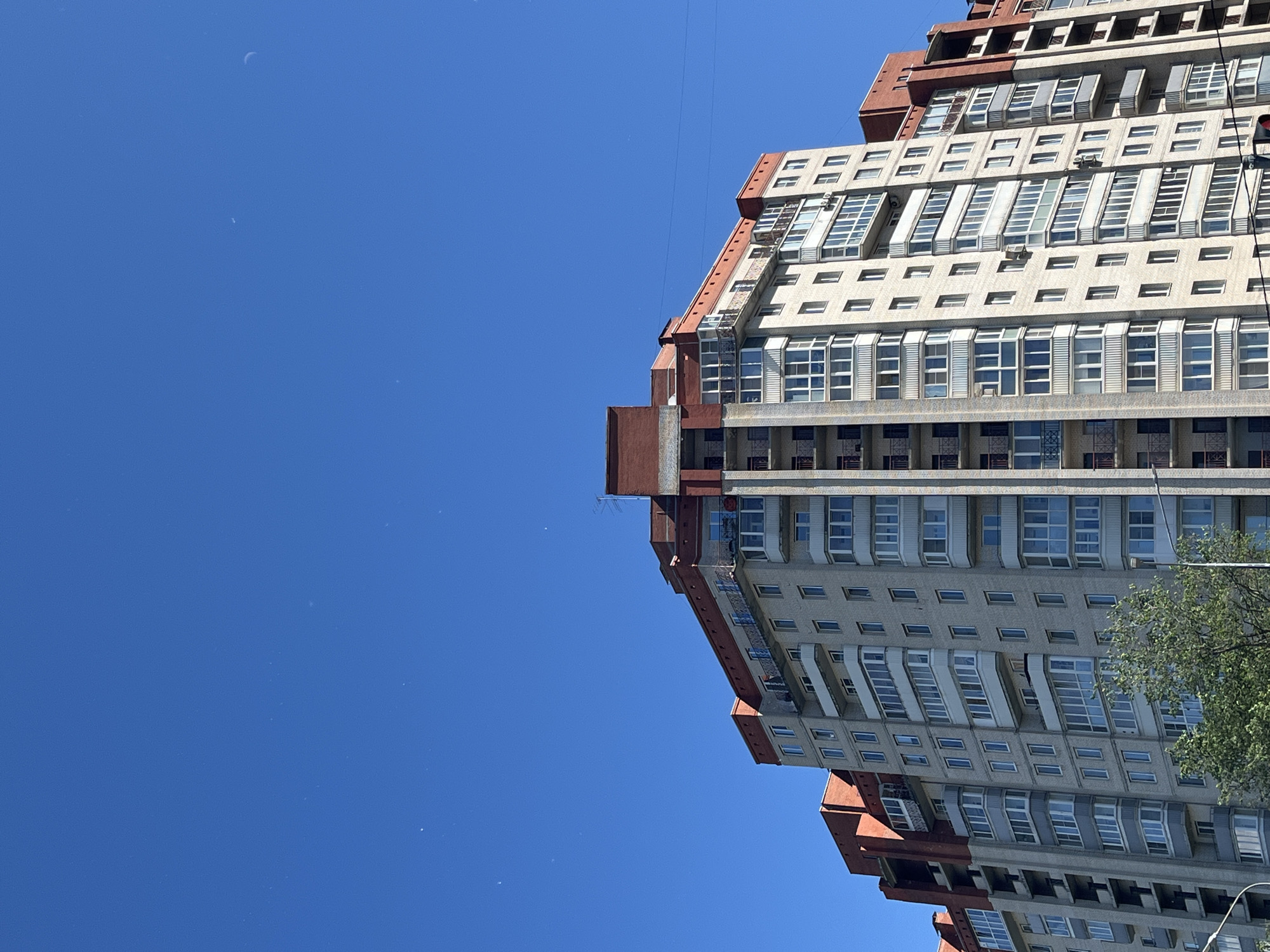 This is a photograph of a modern residential building with a striking, angular design, set against a clear blue sky. The building features a facade of concrete and glass, with numerous windows arranged in geometric patterns. Some windows are recessed, creating a sense of depth, while others have angled coverings that suggest sun protection or architectural detailing. The building's exterior is a mix of light gray concrete and reddish-brown accents on its upper levels and edges, giving it a somewhat Brutalist or post-modern aesthetic.
The perspective of the photograph is unusual, tilted so that the building appears to be leaning or extending upwards from the right side of the frame, with a vast expanse of clear blue sky dominating the left. A thin, dark line, possibly a power line or crane cable, stretches diagonally across the sky. There are a few faint white specks visible in the sky, which could be dust, distant aircraft, or very small clouds. A crescent moon is also faintly visible in the upper left portion of the sky.
To the right of the building, a portion of a lush green tree is visible, its foliage contrasting with the stark lines of the architecture and the uniform blue sky. The bright sky and clear visibility suggest it is daytime, likely midday or early afternoon, with fair weather. The building's numerous windows and balconies indicate it is a residential apartment complex. No people are visible in the image. The context of Saint Petersburg, Russia, suggests this is an urban environment.