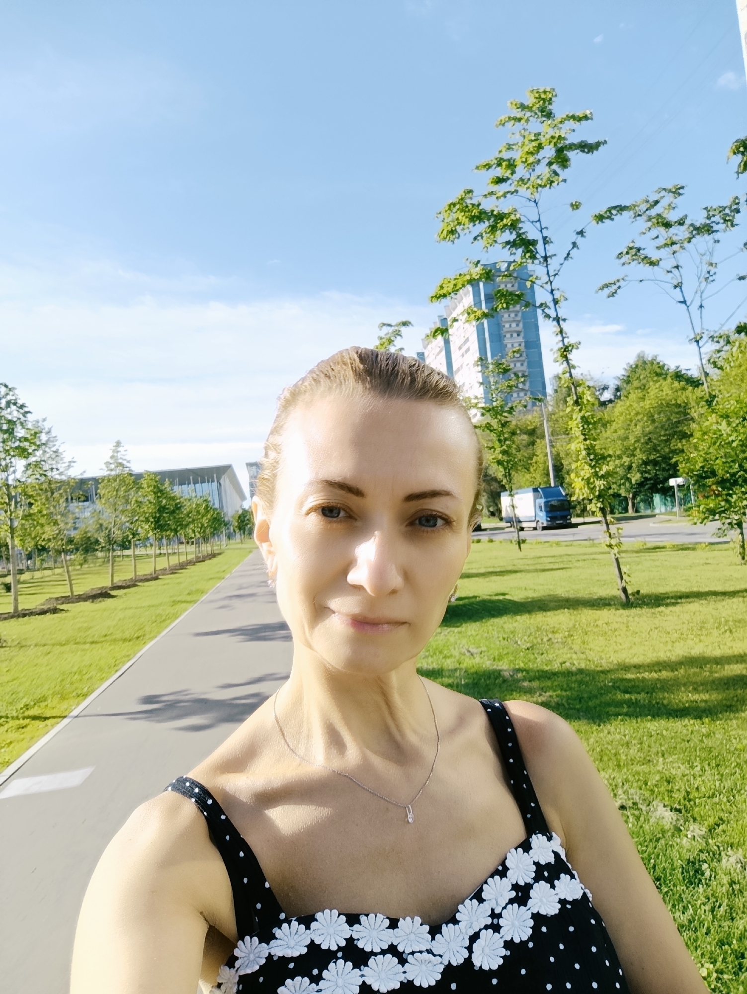 A woman with blonde hair pulled back is taking a selfie outdoors on a sunny day. She is wearing a black dress with white polka dots and a white floral trim. She is standing on a paved path with a grassy area to her right. In the background, there are trees, a tall blue and white building, and a white truck. The environment appears to be a park or a modern urban development. Based on the architecture and vegetation, the location is consistent with a park or green space in Moscow, Russia.