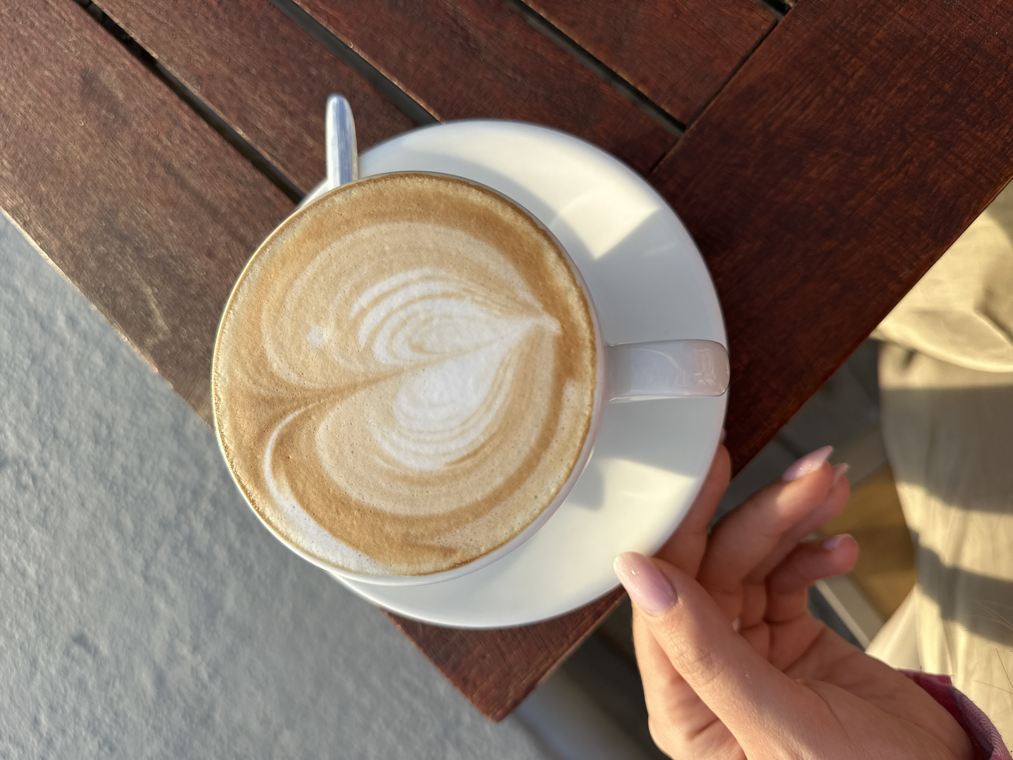A top-down, eye-level close-up shot shows a cup of latte art coffee on a white saucer, on a wooden outdoor table, likely in Bar, Montenegro. A hand is holding the saucer. The coffee art is a heart-like shape. The wooden table is made of slats, and the background includes what appears to be a light-colored concrete surface. The time of day appears to be morning or late afternoon, based on the soft shadows.