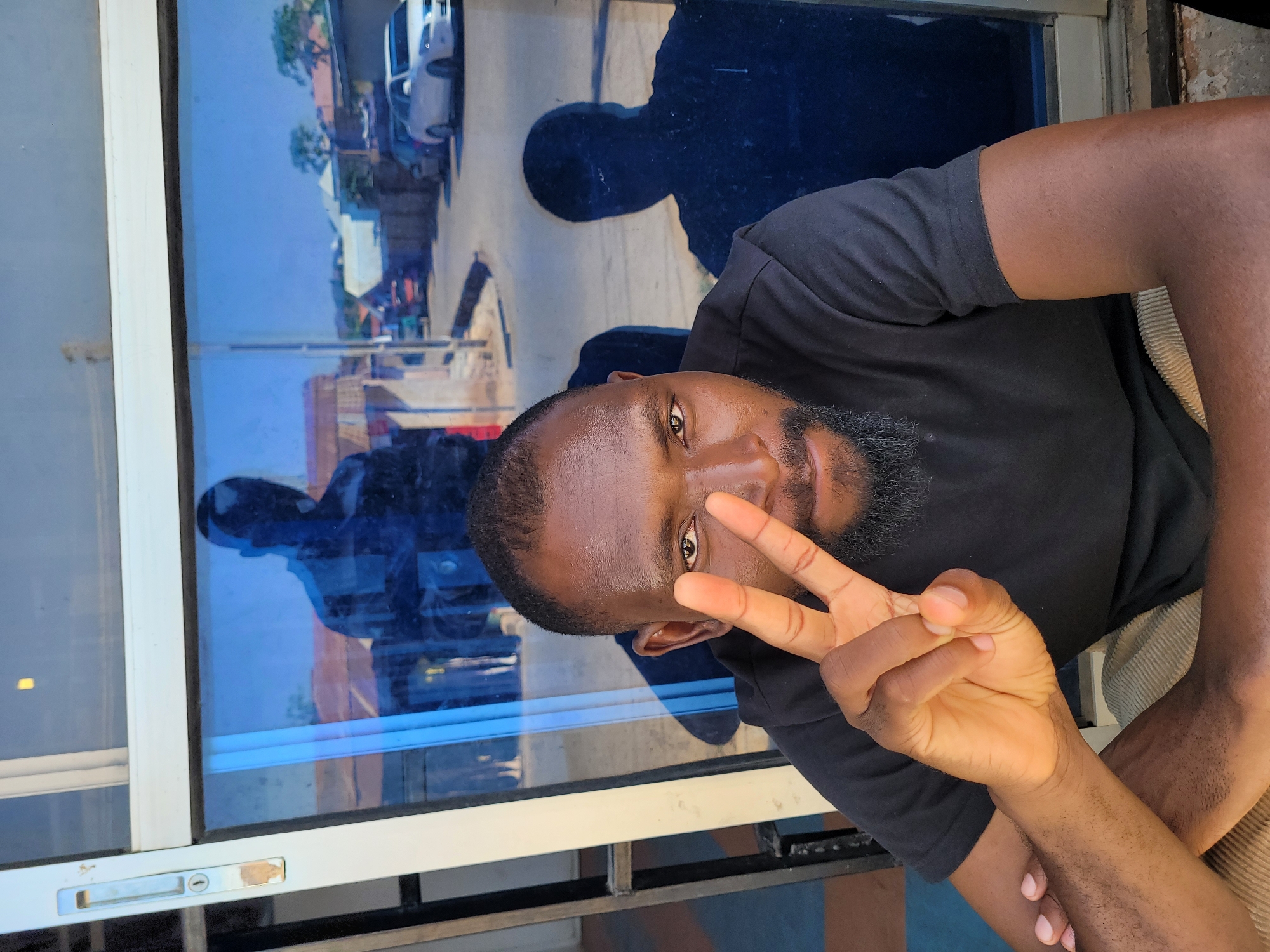 The image is a close-up, slightly angled shot of a dark-skinned man with a beard. He is wearing a black t-shirt and is positioned in front of a window. He is making a peace sign with his right hand. In the reflection of the window, a street scene is visible, with buildings and a white car. The man's shadow can also be seen in the reflection. Based on the limited information, it is difficult to definitively confirm the location, however the style of building and vehicle in the reflection is consistent with the environment of Jos, Nigeria.