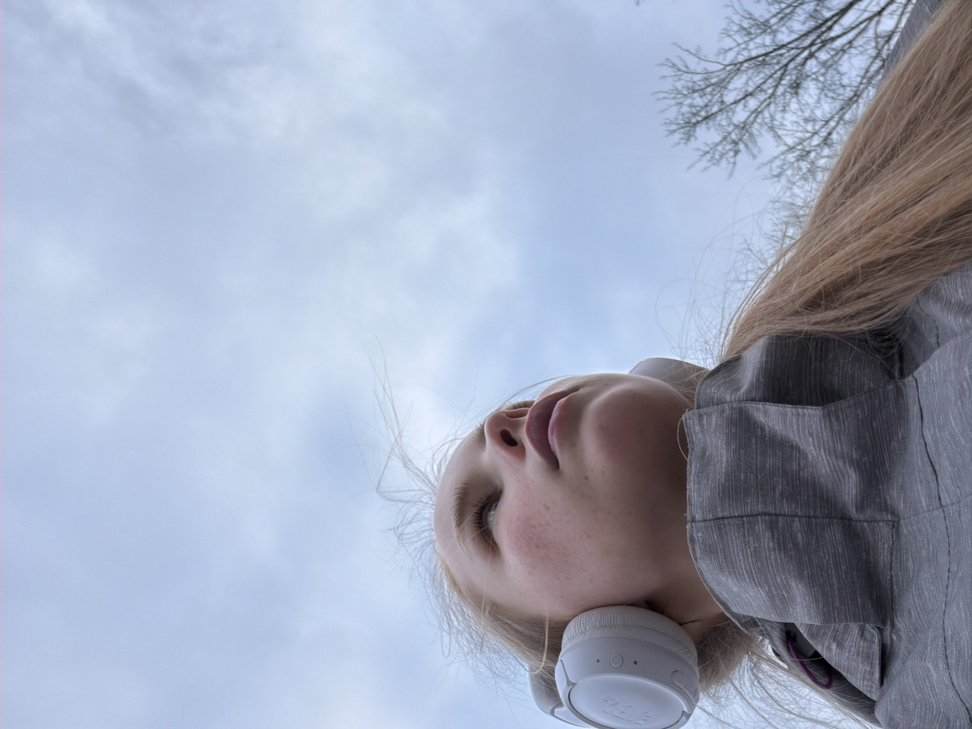 A low-angle shot depicts a young blonde woman wearing white headphones. She is looking upward, with a cloudy sky visible in the background. Bare tree branches are visible in the upper right corner. The woman is wearing a gray textured jacket. The location of Berezniki, Russia is not discernable from the image.